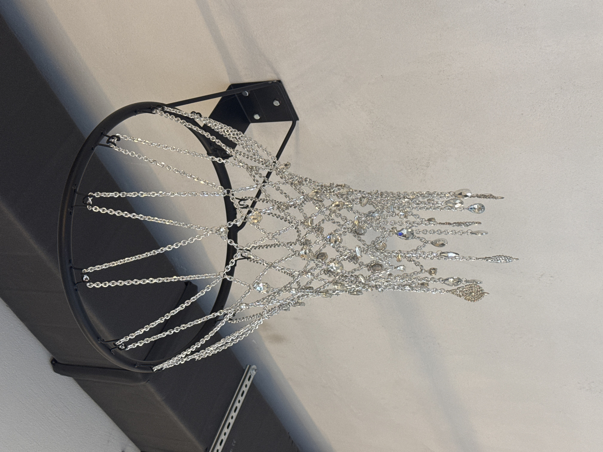 The image shows a basketball hoop with a net made of silver chains and crystals. The hoop is mounted on a wall, and a dark metal beam is visible above it. The wall is painted a light, textured color. The overall impression is of a decorative, artistic interpretation of a basketball hoop. The location of the basketball hoop in the city of Sofia, Bulgaria is not discernible from the image.