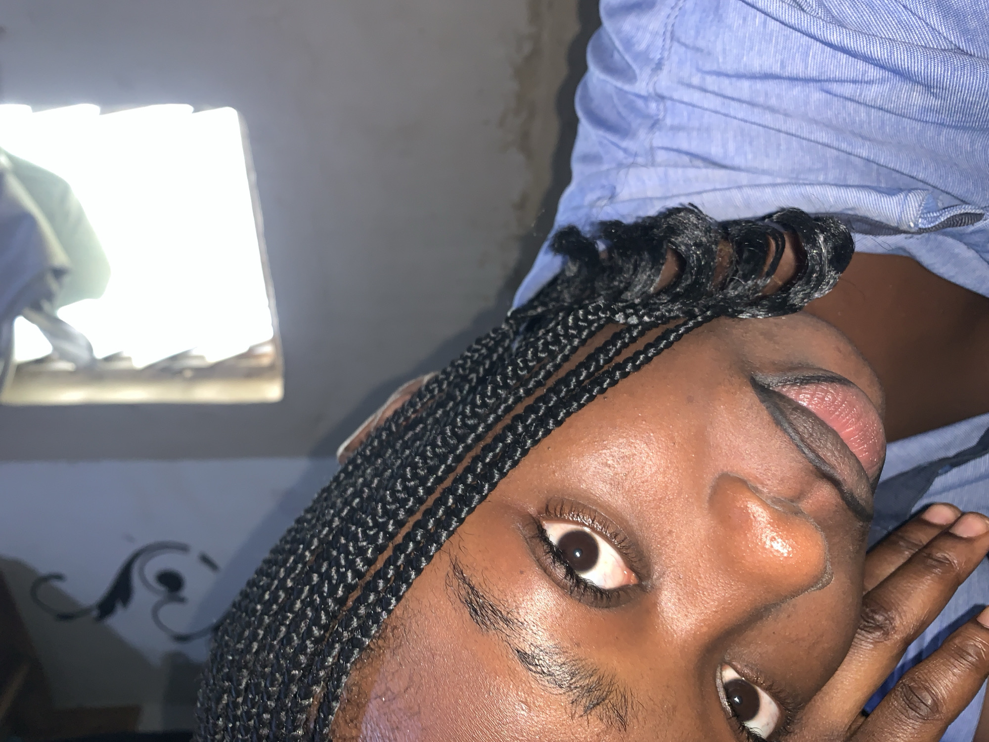 In Rafin Par, Nigeria, a close-up portrait shows a young woman with braided hair resting her head on her hand. She is wearing a light blue collared shirt. The background is dimly lit with a bright rectangular light source visible in the upper right corner. A decorative swirl pattern is visible on the wall to the left.