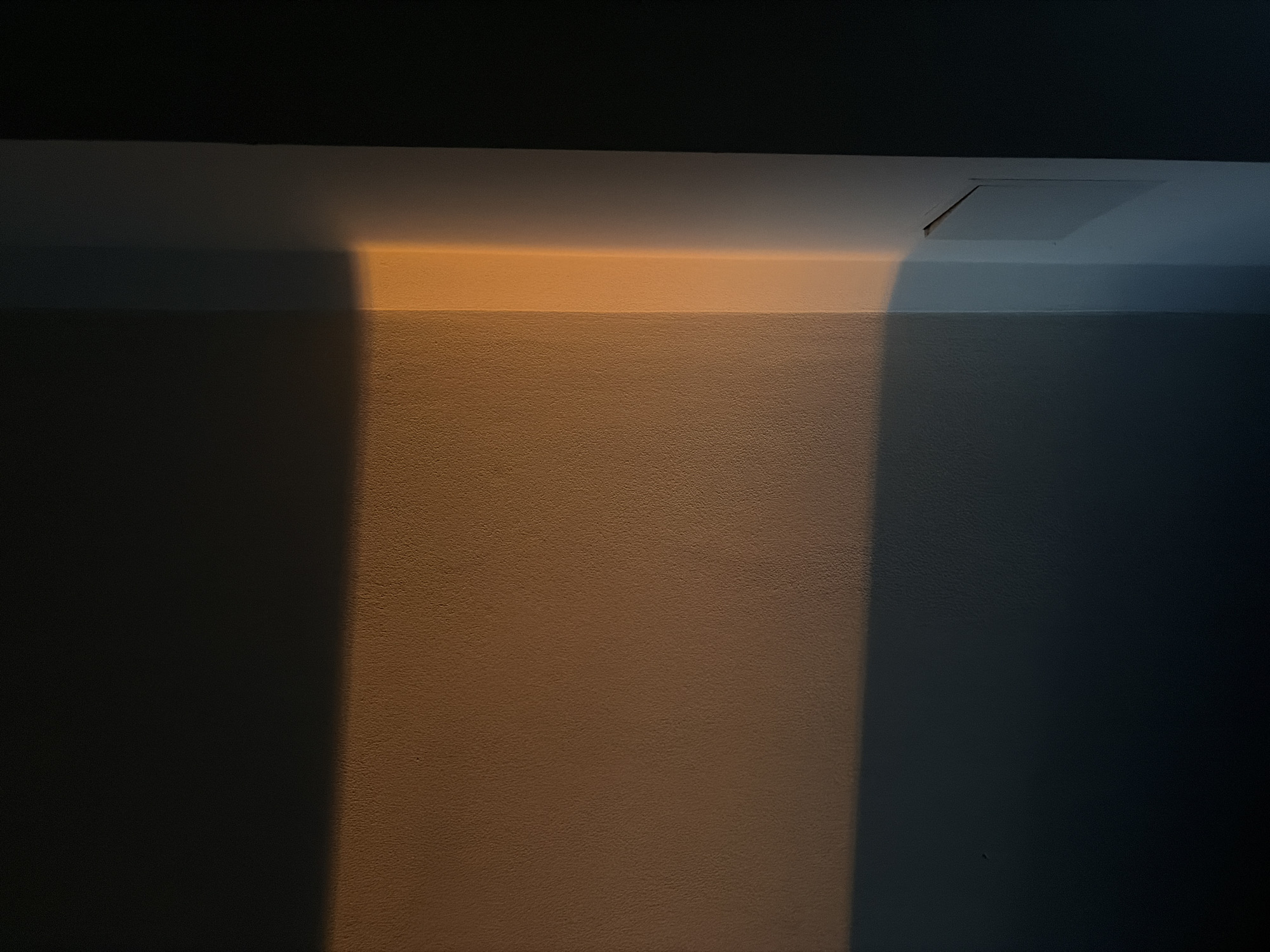 The image depicts a minimalist interior scene focusing on light and shadow play on textured walls. A strong horizontal band of warm, golden light illuminates the upper portion of a wall, transitioning to a darker hue below. To the right, a vertical strip of the same golden light is cast onto an adjacent wall, creating a sharp contrast with the surrounding darker areas and a lighter, possibly white, vertical surface. The textures of the walls are visible, suggesting a matte finish. The scene is devoid of people or overt objects, emphasizing architectural form and the atmospheric effect of lighting. Without specific identifying markers, the location within Nitra, Slovakia, cannot be determined from the image alone.