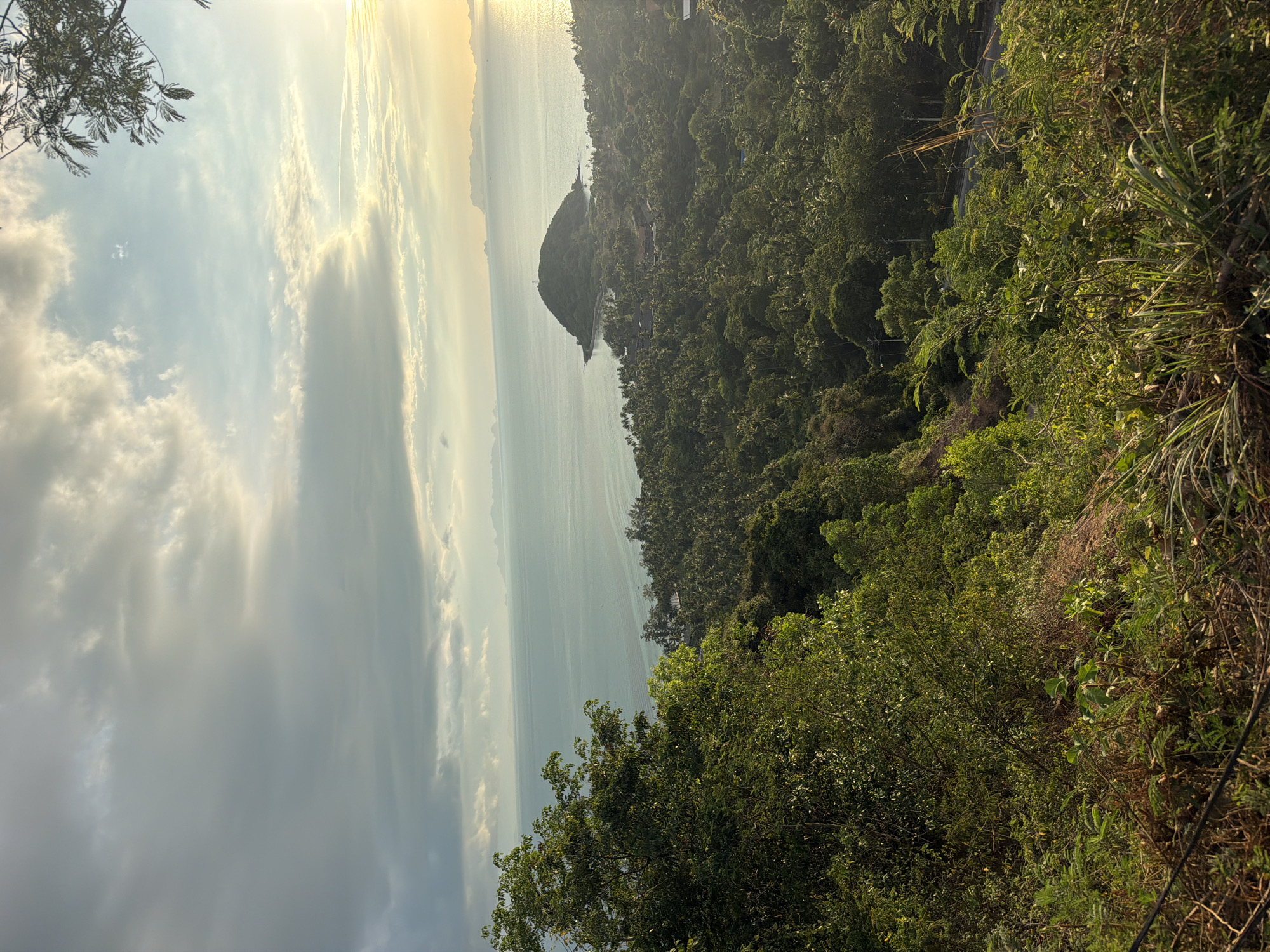 This is an aerial, wide-angle shot of a tropical coastline at sunset. The foreground is dominated by lush, green vegetation, with a steep, brown dirt embankment on the left. A winding road can be seen in the mid-ground, cutting through the dense foliage. Beyond the road, a verdant landscape of palm trees stretches towards the sea. In the distance, a small, conical island with a lighthouse on its peak juts out into the calm, blue water. The horizon is lined with hazy mountains, fading into the warm glow of the setting sun. The sky is a dramatic mix of heavy, grey clouds interspersed with patches of pale blue and streaks of golden light. The overall impression is one of natural beauty and tranquility. The location is specified as Baan Thong Po, Thailand.
