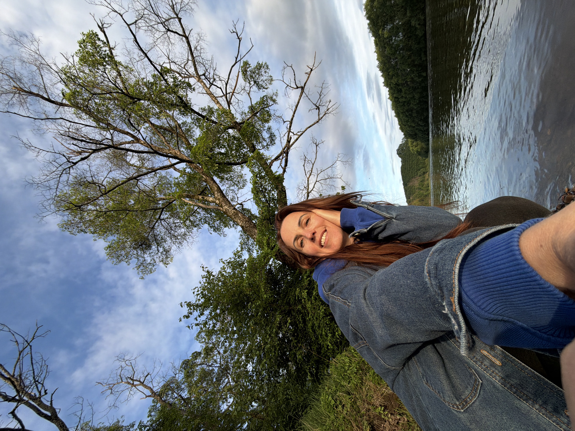The image presents a low-angle outdoor portrait of an adult female taken at the edge of a river in Birobidzhan, Russia. The woman, with long reddish-brown hair, is smiling and looking towards the camera, resting her head on her right hand. She is wearing a blue denim jacket over a bright blue long-sleeved shirt, with a visible blue ribbed cuff on her left wrist. She appears to be sitting or crouching on a rocky and muddy bank at the water's edge, with green grass and foliage visible to her left. The wide river, dark in color with slight ripples, occupies the lower right portion of the frame and extends into the distance. The opposite bank features a dense line of green trees, with undulating, tree-covered terrain visible further back on the left horizon. Above, a large tree with a mix of green leaves and numerous bare branches dominates the upper right quadrant of the sky. The sky displays a mix of light blue and scattered white and grey clouds, suggesting either early morning or late afternoon light.