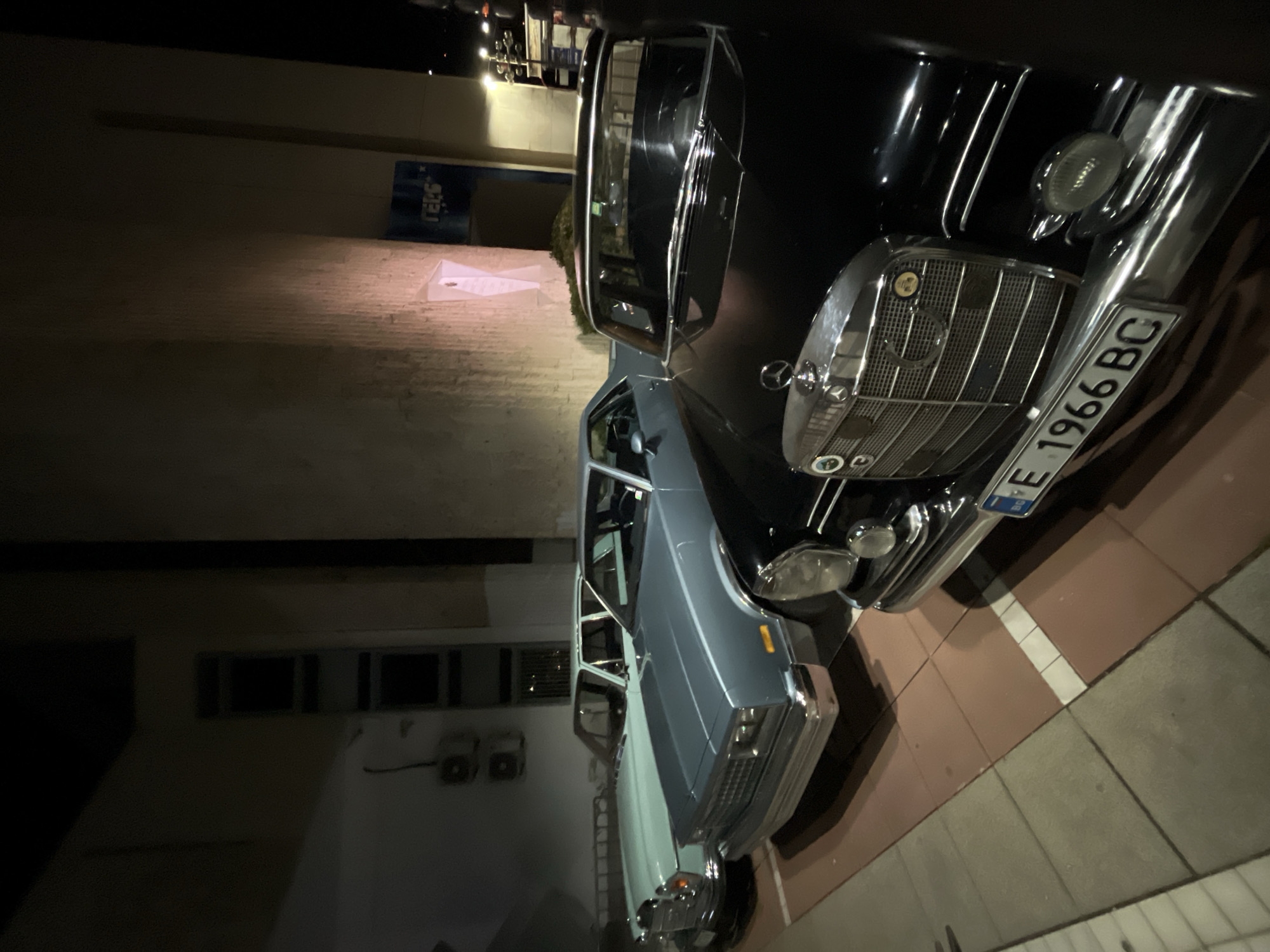 The image captures two vintage cars parked on a tiled surface outdoors, possibly a sidewalk or parking area. One car is dark, potentially black, with a prominent chrome grill, headlights, and the license plate “E 1966 BC”. The second car behind is light blue or gray. The scene seems to be near a building, evidenced by a wall with windows and electrical fittings. In the background, a shop with blue sign can be seen. The presence of a license plate with the “BC” abbreviation suggests the scene is in Bulgaria, and given it's in a city with a shop, a building, and parked cars, it may be in Blagoevgrad, Bulgaria.