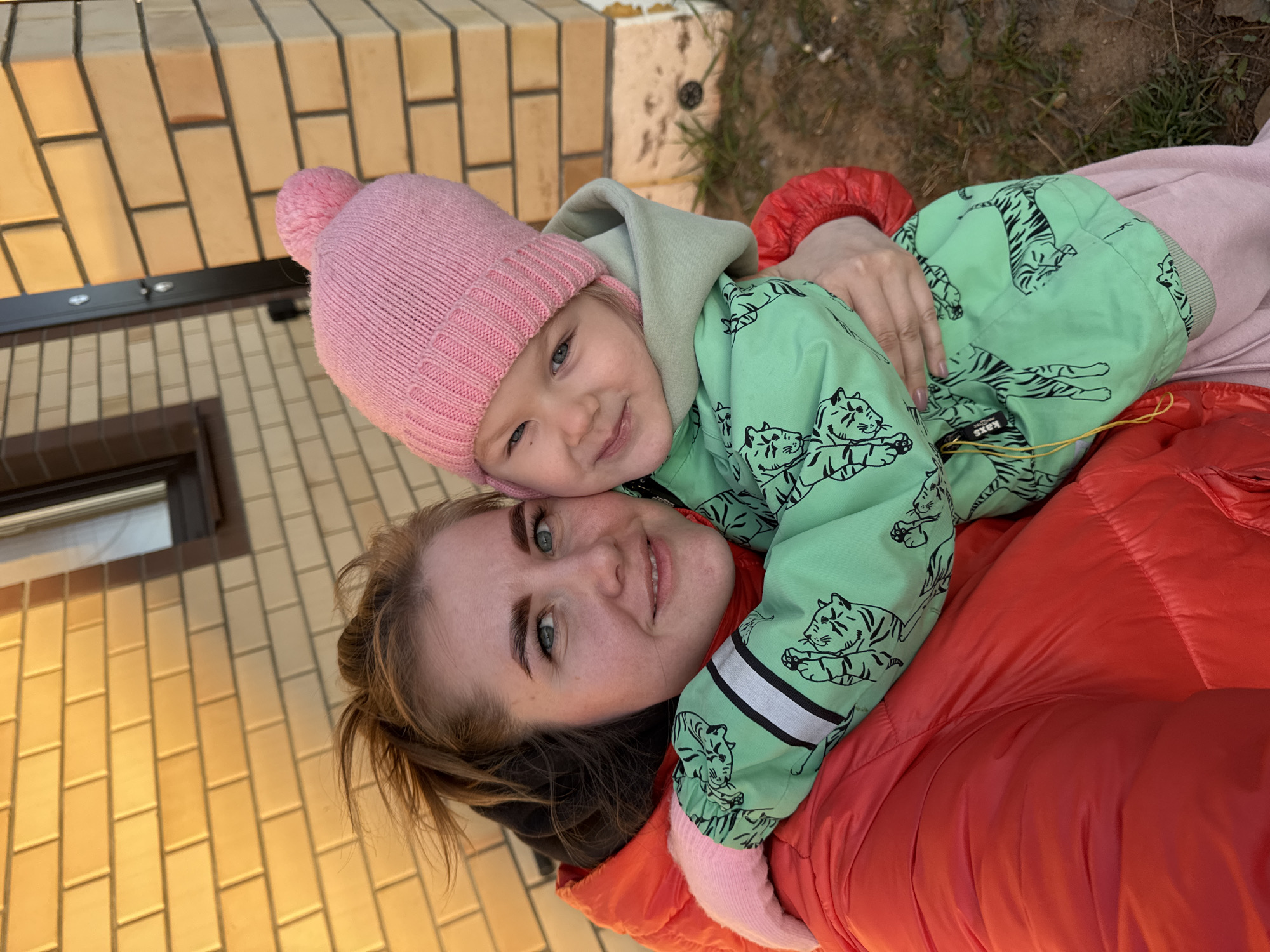 A woman and a young child are lying on the ground in an outdoor setting. The woman, likely an adult, has light skin, fair hair pulled back, and is wearing a bright red puffy jacket. The child, wearing a pink knit hat with a pom-pom and a light green jacket with tiger patterns, is lying on top of the woman. The child is smiling, and their arms are wrapped around the woman's neck.
The background shows a brick building with a window. Based on the location of Ферма, Russia, the environment appears to be a residential area with signs of vegetation in the background.
