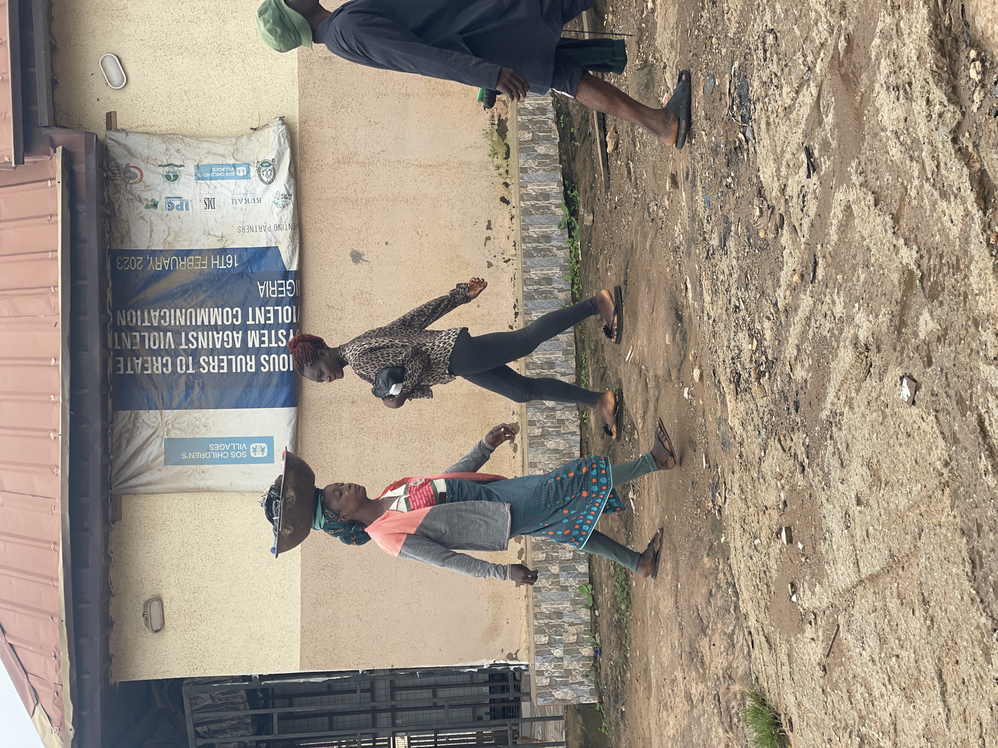 The image depicts a street scene in Kurunduma, Nigeria, with three individuals walking along a dirt and paved road. On the left, a woman wearing a blue and green headscarf balances a large metal basin filled with dark items on her head. She has on a pink and grey jacket over a white shirt with red and green lettering, a blue patterned skirt, and dark green leggings. She is wearing dark sandals. To her right, a younger woman in a leopard-print shirt and black leggings walks with a dark bag in her hand. She is wearing dark sandals and has red hair. Further to the right, a person wearing a dark robe and a green hat walks away from the camera, also in dark sandals. They appear to be carrying a green object. Behind the individuals, a building with a metal roof is visible, adorned with a large banner that reads, "IOUS RULERS TO CREATE SYSTEM AGAINST VIOLENT VIOLENT COMMUNICATION NIGERIA" and "16TH FEBRUARY, 2023". The banner also features logos and text from sponsoring organizations such as "IPS" and "SOS CHILDREN'S VILLAGES". The ground is a mix of dirt, mud, and cracked concrete. Vegetation is sparse, with a few patches of grass visible. The lighting suggests an overcast day.