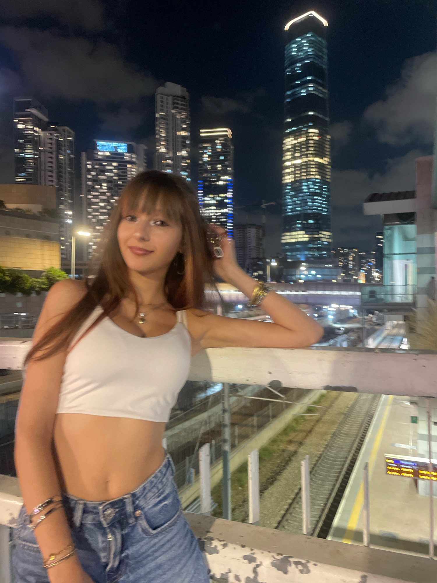 A young woman with long, light brown hair and bangs stands on a bridge at night. She is wearing a white crop top and blue denim shorts. Her left arm is resting on the railing, and her right hand is near her head. Behind her, a city skyline is visible with numerous illuminated skyscrapers. The image appears to be taken in Tel Aviv, Israel, likely near a train station, as train tracks and a platform are visible below the bridge. The sky is dark with clouds, and the city lights create a vibrant backdrop.