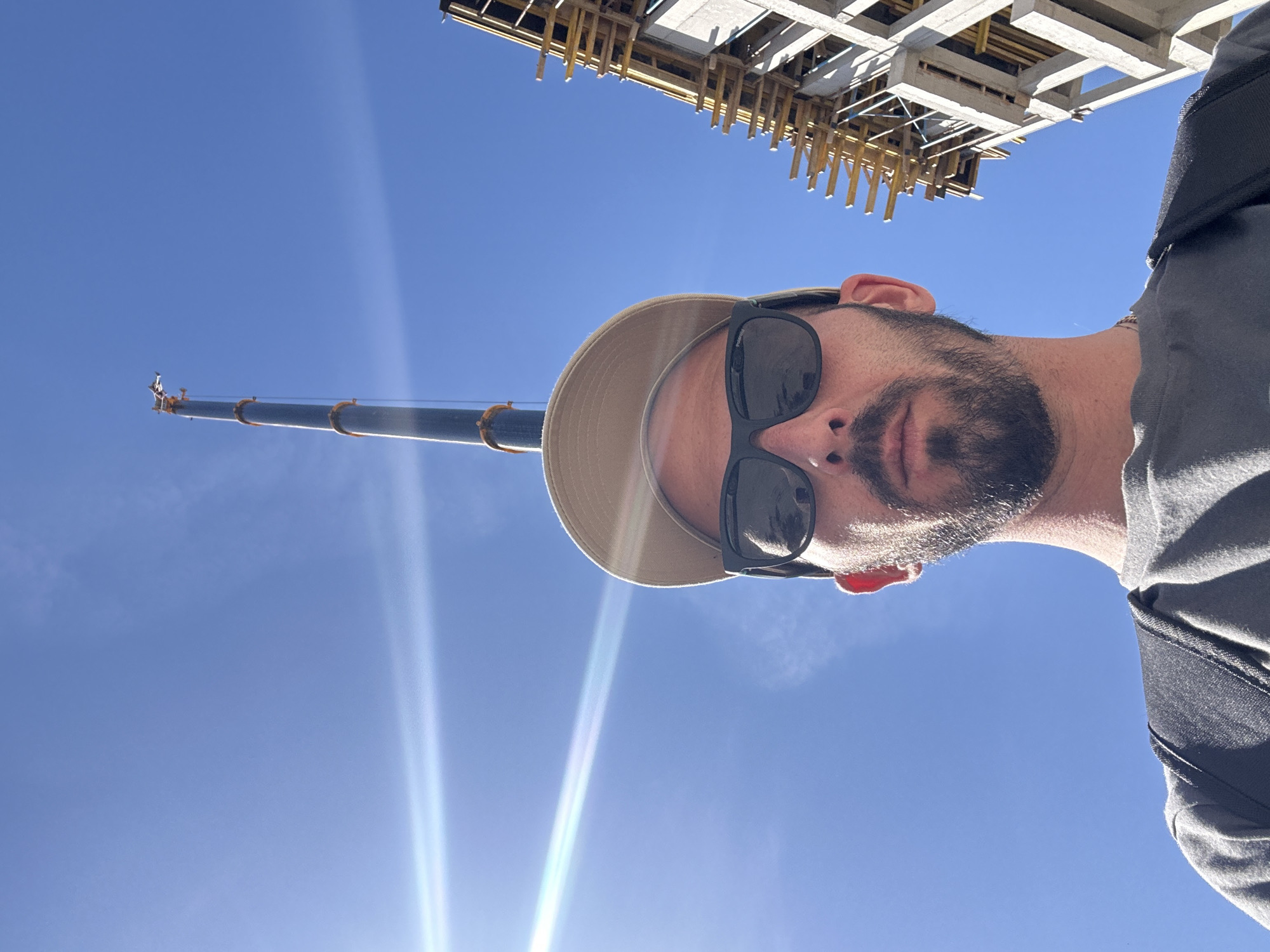 A low-angle selfie, taken outdoors in Budva, Montenegro, captures a man looking upwards towards a bright, clear blue sky. The man is positioned in the foreground, appearing inverted due to the camera's orientation. He wears black sunglasses, a light beige wide-brimmed hat, and a grey shirt or t-shirt, with a dark beard covering his chin and jawline. The brand "Polaroid" is faintly visible on the temple of his sunglasses.
Against the vibrant sky, a tall, dark-colored construction crane's arm extends from the left side of the frame, stretching horizontally. Its far end, or jib, is visible, indicating its substantial height. In the upper right corner, a concrete and wooden scaffolding structure, typical of a building under construction, can be seen, suggesting an active construction site.
The scene is brightly illuminated by natural sunlight, resulting in prominent lens flares in the lower part of the image and reflections off the crane's surface. The clear sky indicates a sunny day. The overall impression is a casual snapshot taken at a construction site.