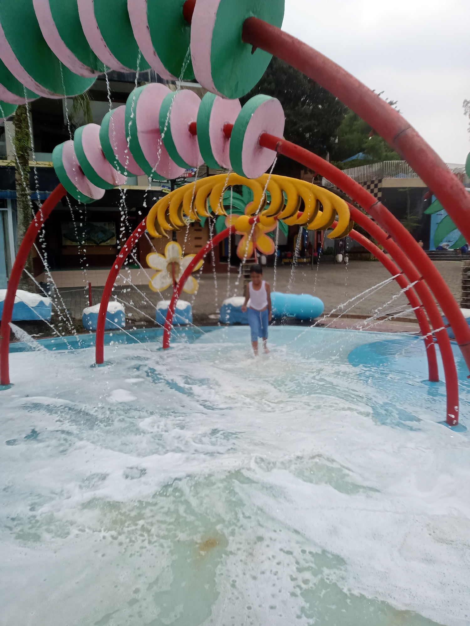 The image depicts a water play area in a public place, possibly a water park or resort, located in Baru, Indonesia. A young child, wearing a white tank top and blue shorts, is standing in shallow, foamy water, with water spraying around them from overhead structures. These structures feature a series of colorful, oversized discs in pink and green, resembling an abacus, and a yellow, banana-shaped arch. Red support beams frame the play area, and in the background, there are more water features, buildings, and greenery. The ground appears to be paved, and the water is clear and blue in some areas, transitioning to foamy white where the water is actively spraying. The overall atmosphere is one of recreation and fun.