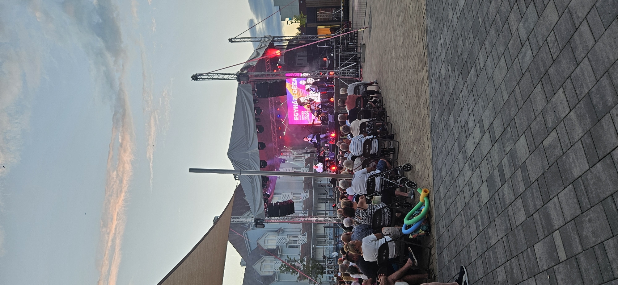 The image captures an outdoor concert performance in the city of Hévíz, Hungary. A stage is set up with musicians performing, illuminated by stage lights and a large screen displaying "EGYHÁZI GÉZA". A crowd of people, mostly older adults, are seated in rows facing the stage. Some individuals are using walkers or strollers. In the foreground, a paved area leads to the seating, with some colorful inflatable toys on the ground. The background shows buildings, trees, and a cloudy sky with birds flying. The overall atmosphere appears to be a relaxed evening event.