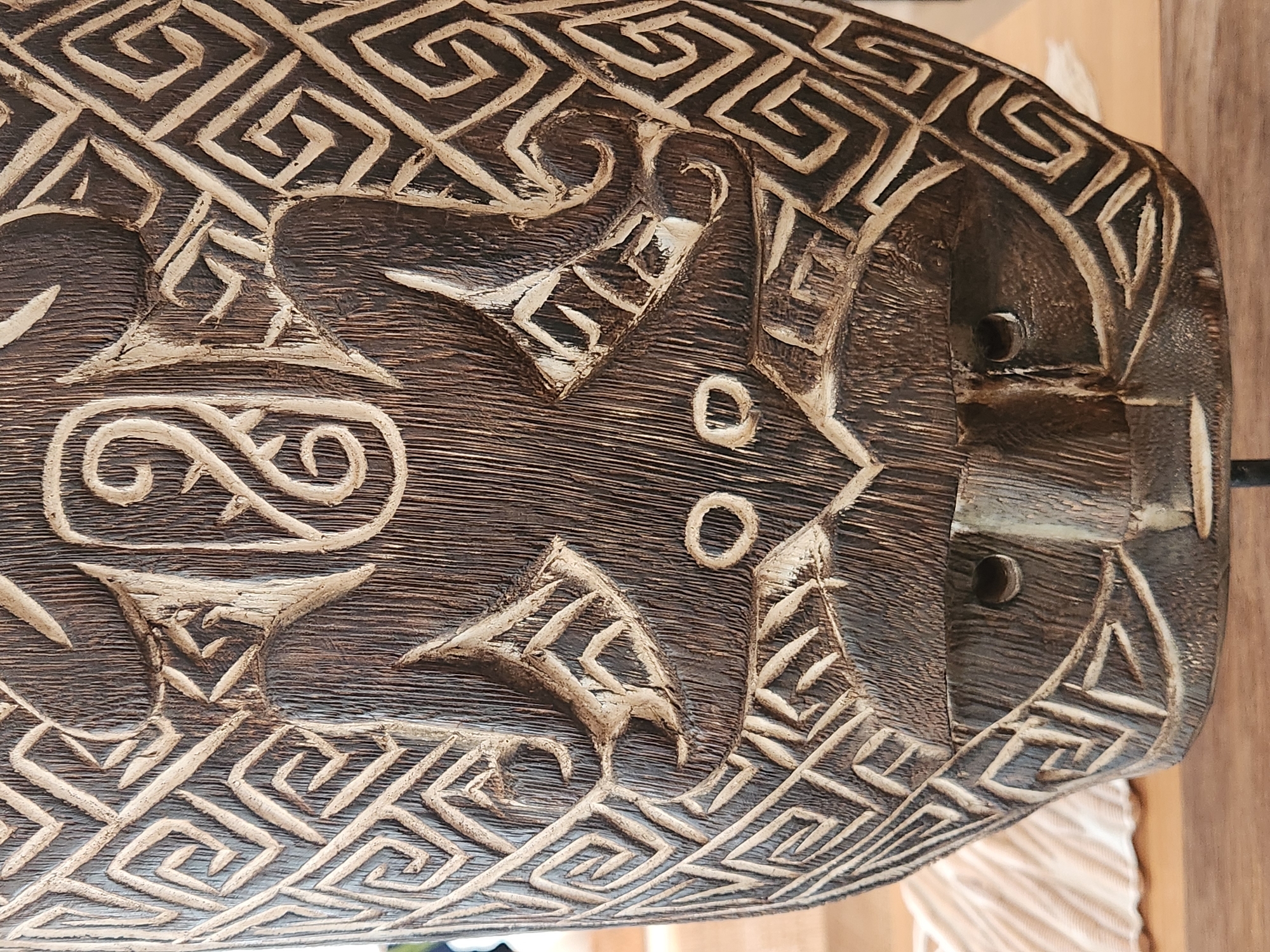 The image displays a close-up, vertical shot of a carved wooden mask. The mask is dark brown and features intricate geometric patterns, primarily composed of interlocking squares and spirals, carved into its surface. In the center of the mask, there is a stylized representation of a face with two circular eye indentations and a prominent, wide mouth area. Above the eyes, there are two abstract, human-like figures with outstretched limbs carved into the mask. The texture of the wood grain is visible throughout, indicating a handcrafted item. The mask is positioned upright, likely on display. The background is out of focus but suggests an indoor setting with a light-colored, textured surface to the left of the mask and a wooden surface below. There is no discernible location information within the image itself.