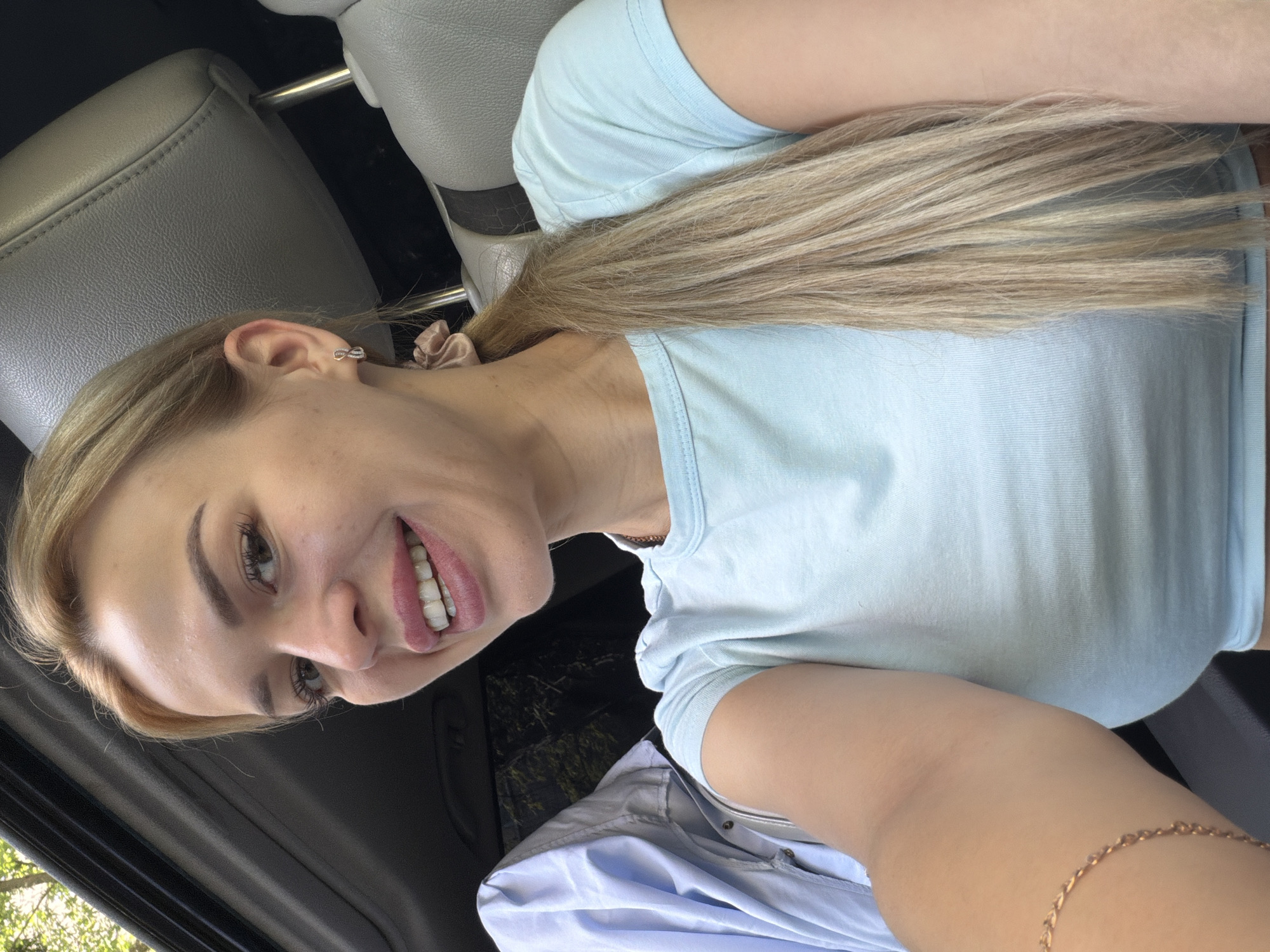 In Kyiv, Ukraine, a young blonde woman with fair skin is taking a selfie inside a car. She is wearing a light blue t-shirt and a delicate gold bracelet on her left arm. Her blonde hair is pulled back into a ponytail. Her smile reveals white teeth and pink lipstick. The car interior, with its gray leather seats and sunroof, is visible. A light blue garment is hanging on the back of the driver's seat. The overall scene suggests a casual, everyday moment.