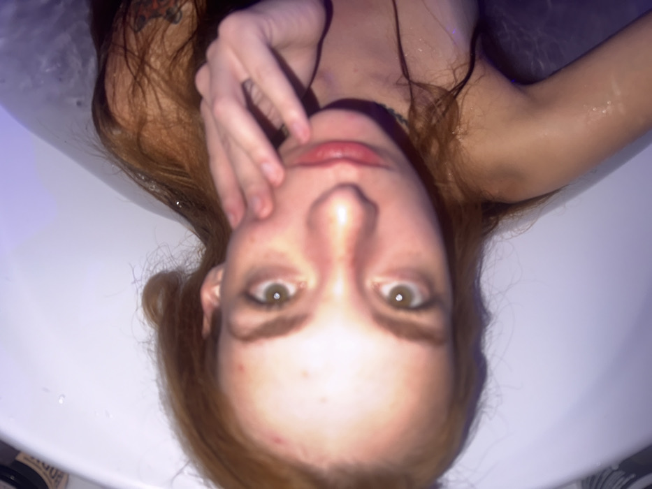 An overhead-angle image depicts an individual with reddish-brown hair partially submerged in a white bathtub. The scene is indicated as occurring in the city of Khimki, Russia. The individual's head, shoulders, and upper torso are visible. Their skin shows multiple small blemishes on the forehead and cheeks. The individual's left hand is raised, with fingers touching the lower left side of their face. Their eyes are open and directed at the camera. A dark, possibly tattooed, marking is visible on the right upper arm/shoulder area. A thin, dark necklace or choker is worn around the neck. The bathtub contains water with scattered foam/bubbles on the surface. A dark cylindrical object, resembling a container, is partially visible on the tub's edge in the upper left. The environment is illuminated by what appears to be artificial light, casting a purple ambient glow on the water and background.