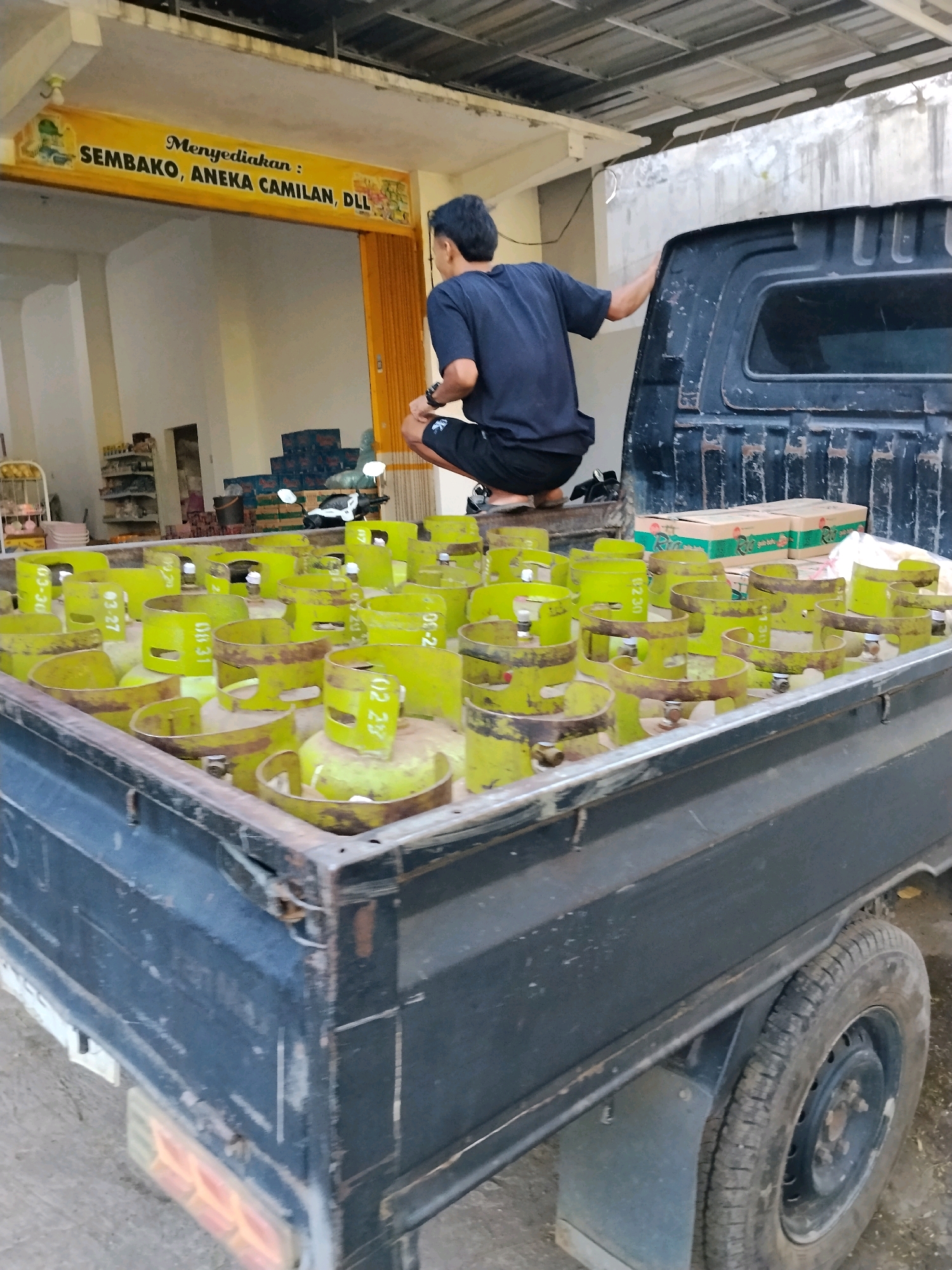 The image depicts a pickup truck loaded with numerous green propane tanks, parked in front of a building in Panggungan, Indonesia. A young man in a dark blue t-shirt and black shorts is crouched on the edge of the truck bed, facing away from the camera. He appears to be looking into the distance, with his left hand resting on the truck's cabin. The truck bed is filled with propane tanks, some of which are labeled with numbers. In the background, a yellow banner above a doorway reads "Menyediakan : SEMBAKO, ANEKA CAMILAN, DLL" (Provides: Groceries, Assorted Snacks, etc.). The building has a metal roof and an orange accordion-style door. Several stacks of cardboard boxes are visible inside the building, along with shelves stocked with various items. The overall scene suggests a commercial setting, possibly a small grocery or convenience store, where propane tanks are being delivered or prepared for distribution. The ground appears to be paved with concrete or asphalt.