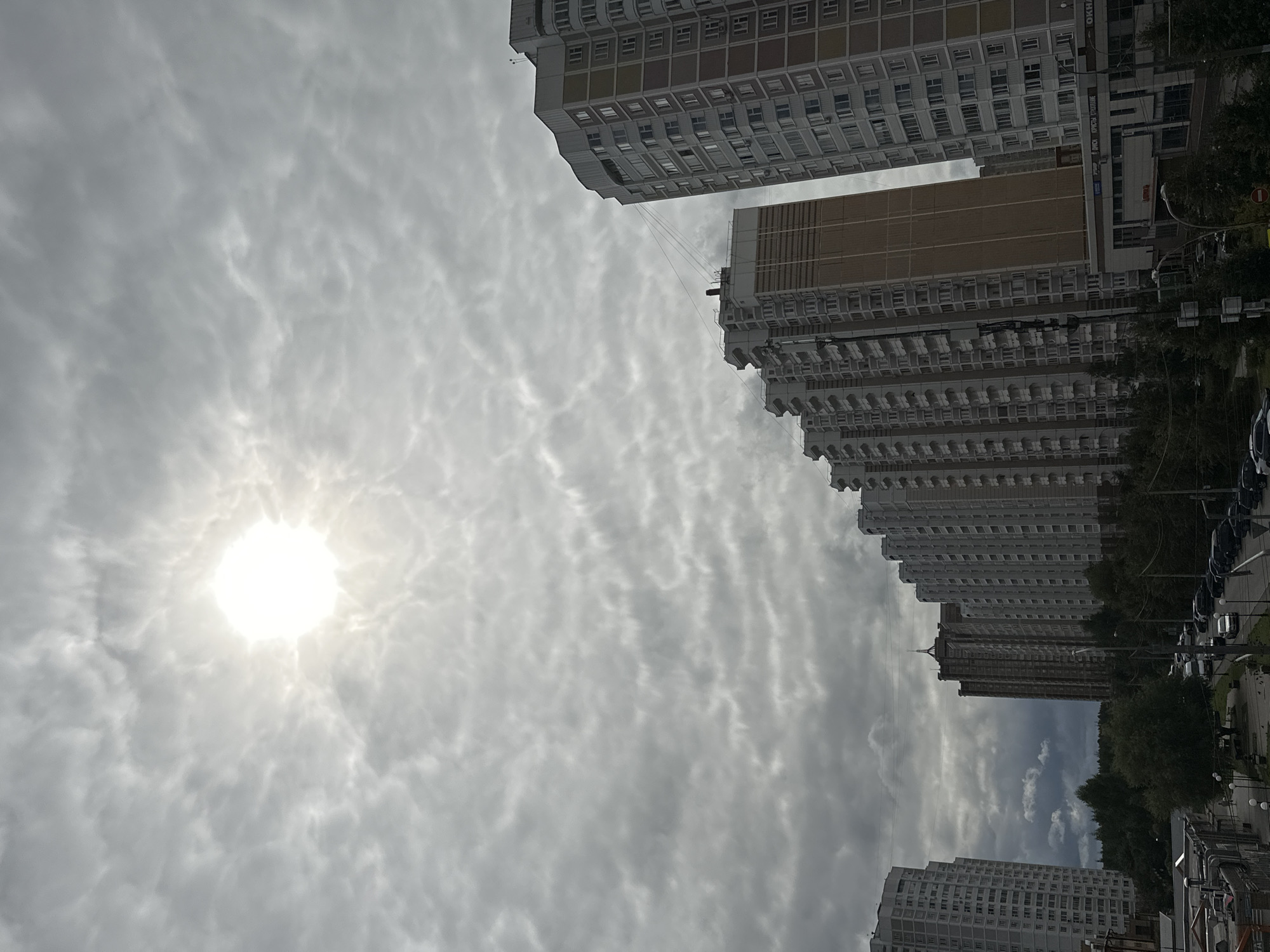 A daytime, low-angle exterior shot captures a cloudy sky with the sun shining through the overcast in Podolsk, Russia. The sky is filled with textured, grey clouds that form wavy patterns, with the bright sun creating a halo effect. In the foreground, a line of multi-story apartment buildings stretches across the frame, their facades predominantly grey, white, and beige, with many windows visible. Some buildings feature balconies. Below the buildings, a street is lined with trees and parked cars, indicating a residential area. The scene is static, with no people or discernible actions taking place.