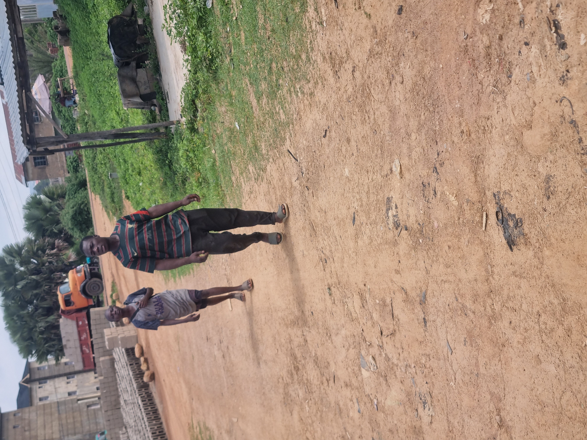 Two young males are walking on a dirt road, in what appears to be a semi-urban or rural setting. The male in the foreground is wearing a striped t-shirt with red, green, and black stripes, and dark pants. He is walking towards the viewer. The second male, slightly behind and to the left, is wearing a gray t-shirt with writing on it and dark shorts. He is also walking in the same direction as the first male.
In the background, to the left, a pile of what appears to be building bricks or blocks is stacked. Further back, a large orange truck with a red flatbed is parked. A building with multiple stories and concrete construction is visible behind the bricks. Palm trees and other green vegetation are also present in the background, suggesting a warm climate. A rustic shelter with a corrugated metal roof is located in the mid-ground to the right. Underneath the shelter, a table and chairs are set up, with an umbrella providing shade.
The ground is a dusty, reddish-brown dirt, with scattered debris. The overall impression is of a construction site or an area undergoing development. The lighting suggests it is daytime, possibly overcast due to the diffused light and lack of strong shadows. There is no specific visual information that definitively places this scene in Jos, Nigeria, but the general environment with the type of buildings, vegetation, and the presence of construction is consistent with many areas in Nigeria.