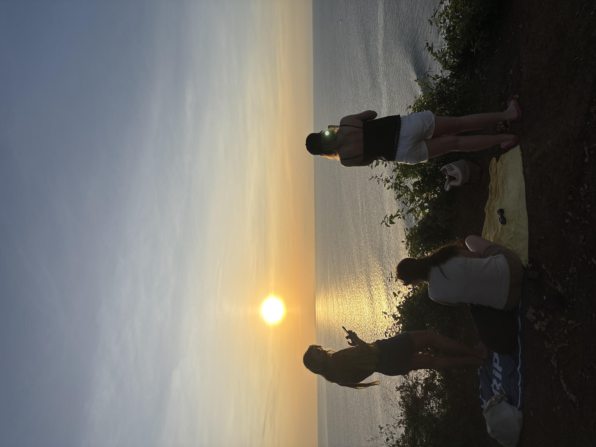 The image captures a scenic view at Kuta Selatan, Indonesia, featuring three women enjoying a sunset. The setting appears to be a cliff overlooking the ocean. The woman nearest the top of the image is wearing a black top and white shorts. She has a hat on and appears to be looking towards the ocean. Below her, two other women are sitting on a mat or blanket placed on the ground. One of them is wearing a light-colored shirt and the other is wearing a mustard shirt while holding a mobile phone. The sun is setting over the ocean, casting a golden hue across the sky and water.