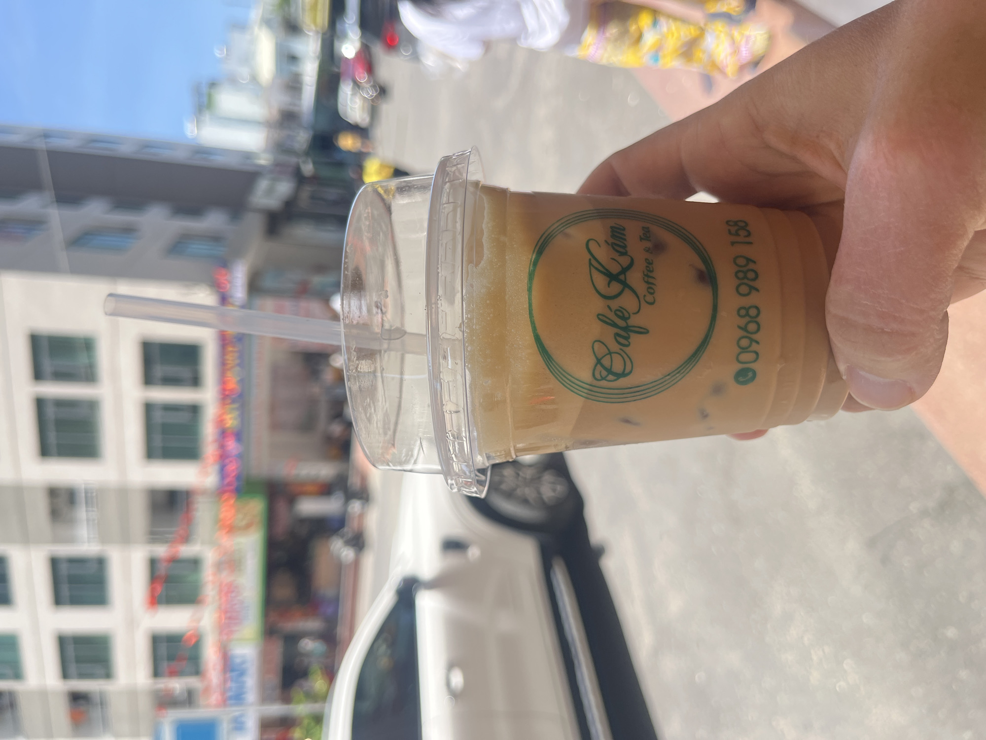 A forensic examination of the image reveals a close-up perspective of a light-skinned hand holding a clear plastic cup filled with an iced coffee beverage on a bright, sunny day. The cup contains a light brown liquid, ice cubes, and small dark particulates. A clear plastic lid with a straw opening covers the cup, and a clear plastic straw extends from it. A green circular logo is printed on the cup, displaying "Cafe Lám" in a script font, followed by "Coffee & Tea" in a smaller sans-serif font. Below the logo, the phone number "0968 989 158" is printed in green.
The background is significantly blurred, indicating a shallow depth of field, and depicts an urban street scene in what is identified as Bac Nha Trang Ward, Vietnam. Prominent in the blurred midground on the left is a multi-story building with a light-colored facade and multiple windows, some reflecting a green hue. Red banners or decorations are visible strung across the street or between buildings in the mid-distance.
A white SUV or car is partially visible on the left side of the street, its front and side in the midground. Numerous other blurred vehicles, including what appear to be motorbikes and cars, are present further down the street, with a red car's taillight discernible on the right. A person wearing a white t-shirt and patterned yellow or floral pants is partially visible on the far right, walking away from the camera. The ground appears to be paved, and the strong sunlight casts clear shadows, indicating outdoor conditions.