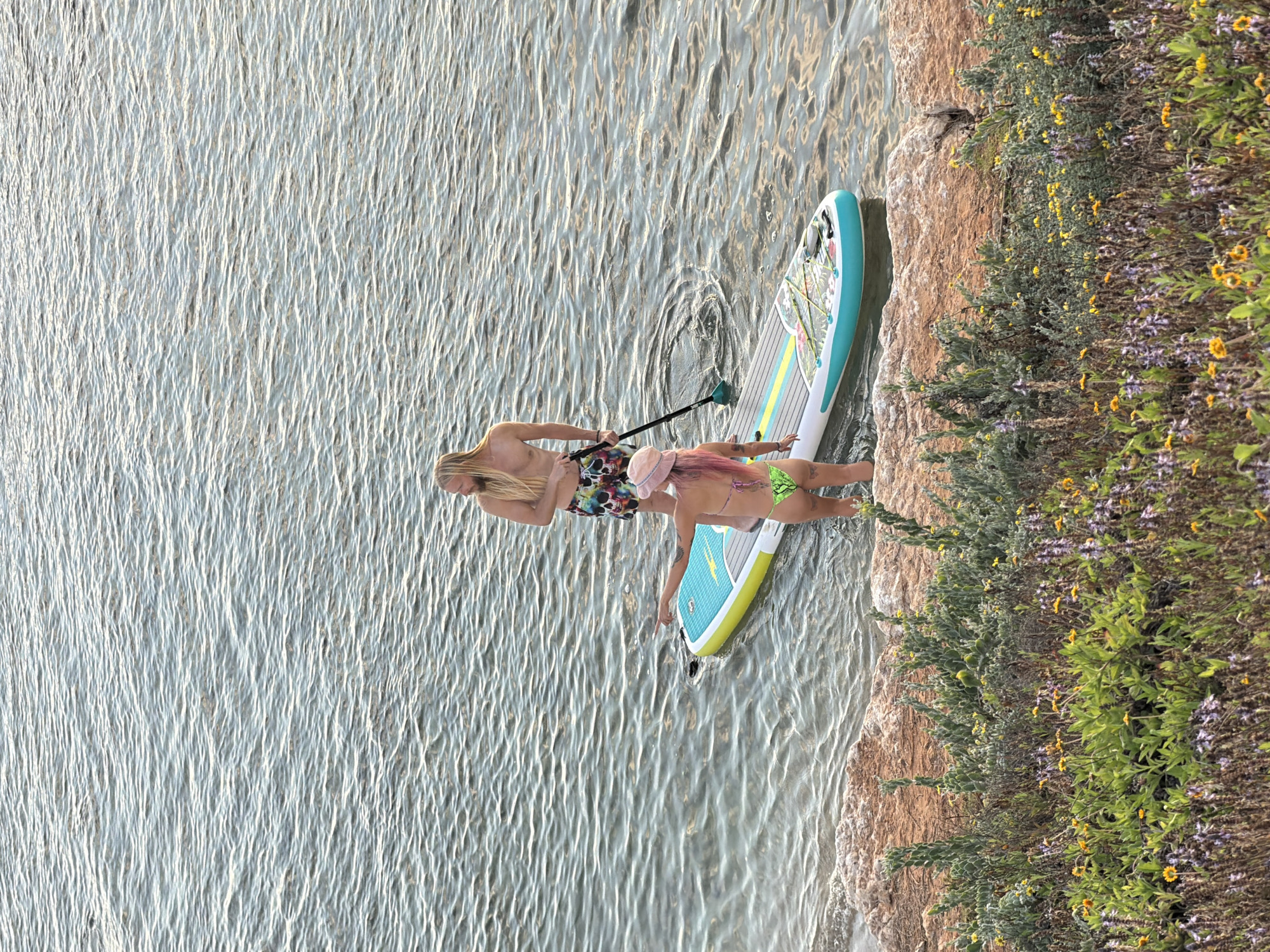 In Tel-Aviv, Israel, a couple enjoys a paddleboarding adventure on a body of water, likely the sea, given the location context. The scene is bathed in the soft light of what appears to be late afternoon or early morning. The water is calm, with gentle ripples reflecting the sky. The couple is positioned on a single paddleboard, its colorful design a vibrant contrast against the water. The man, with long blond hair, is partially visible, holding a paddle. Beside him, a woman with pink-tinted hair is wearing a green bikini and a pink hat. The paddleboard is close to a rocky shore adorned with lush green vegetation and blooming wildflowers, some yellow and others a soft purple, adding a natural, scenic element to the setting. The overall mood is relaxed and idyllic, suggesting a pleasant recreational outing. No visible text is present in the image.