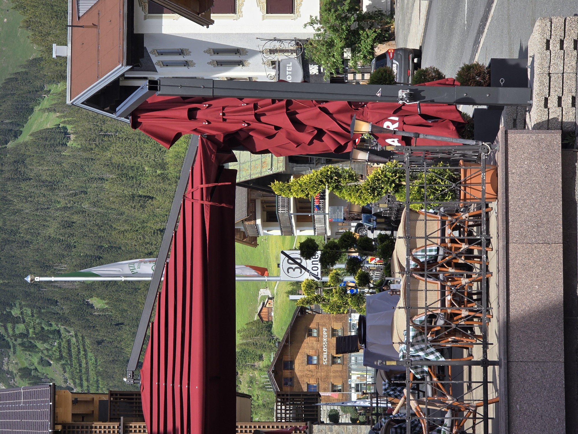 An exterior view captures a sunlit scene in Lech, Austria, likely during warmer months, as indicated by the lush green mountainside dotted with trees and terraced fields in the background, and the presence of outdoor seating at a restaurant.
In the foreground, a red retractable awning shades an outdoor dining area furnished with tables covered in checkered cloths and chairs made of wicker and wood. A dark metal railing encloses this space. Two potted evergreen trees stand to the right of the railing.
Behind the outdoor seating, several buildings are visible. The closest one on the left is a wooden structure with a dark tiled roof. Further back, a larger wooden building features multiple windows and the name "SCHLOSSKOPF" prominently displayed. To its right, a white building with a red roof sports architectural details and balconies. A sign for "HOTEL" is affixed to its facade.
A white flagpole with a green and white flag stands between the wooden buildings. To the right of the flag, a circular "30 ZONE" speed limit sign is posted. A dark gray car is parked on the asphalt road in the middle ground to the right. The overall atmosphere is tranquil, suggesting a typical day in a mountain resort town.