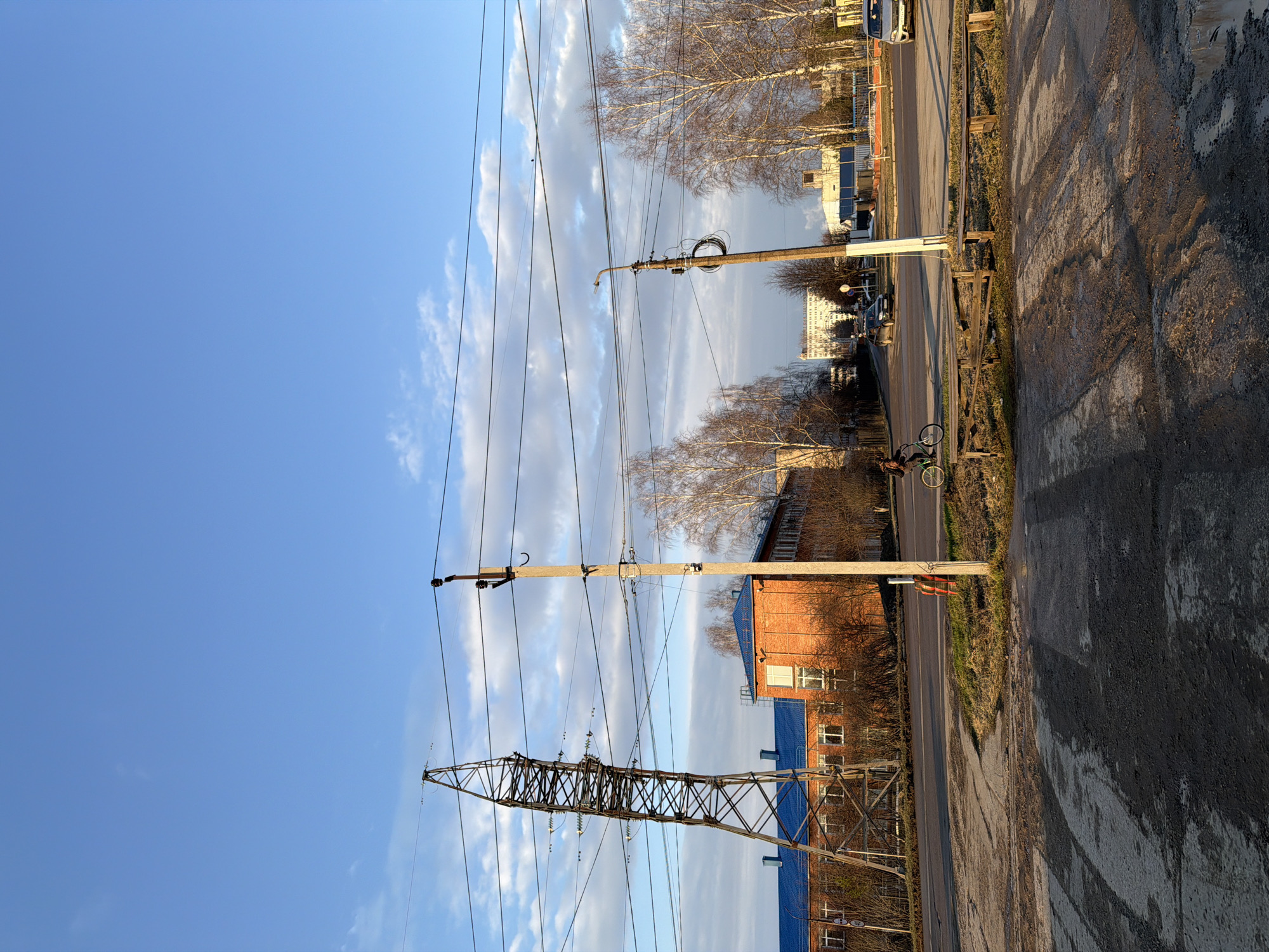 The image captures a view in Chaikovsky, Russia. Power lines stretch across the frame, supported by a metal lattice tower and two concrete poles. A red brick building with a blue roof is visible on the right side of the image. A paved road runs parallel to the buildings and power lines. Bare trees are present in the background. The sky is predominantly blue with scattered clouds. The ground surface appears to be asphalt with some patches of moisture. A bicycle is parked near the side of the road.
