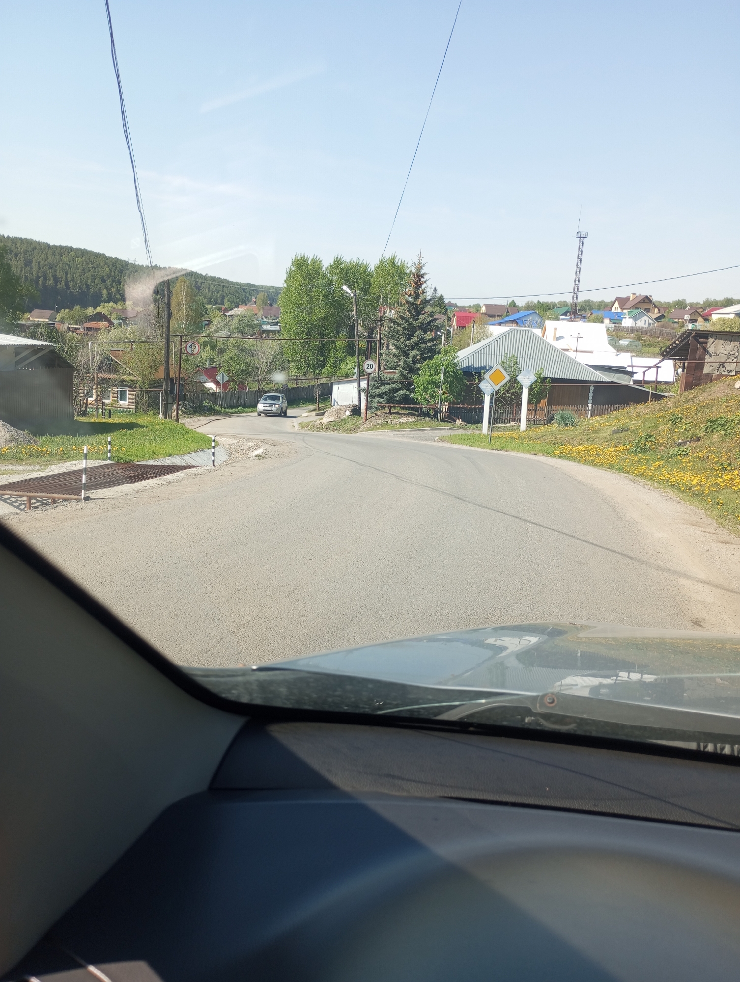 The image, taken from the interior of a vehicle, presents a view of a road in the city of Кодинка, Russia. The road curves slightly to the right, and the asphalt surface appears well-maintained. In the distance, a silver car is visible. To the left of the road, there is a cattle grate and a structure resembling a covered shed. On the right side of the road, there is a yellow diamond-shaped road sign. Beyond the road and adjacent to the buildings, a field of yellow flowers stretches across the landscape. The surrounding environment includes scattered houses and trees, with forested hills in the background. Overhead, power lines extend across the sky.