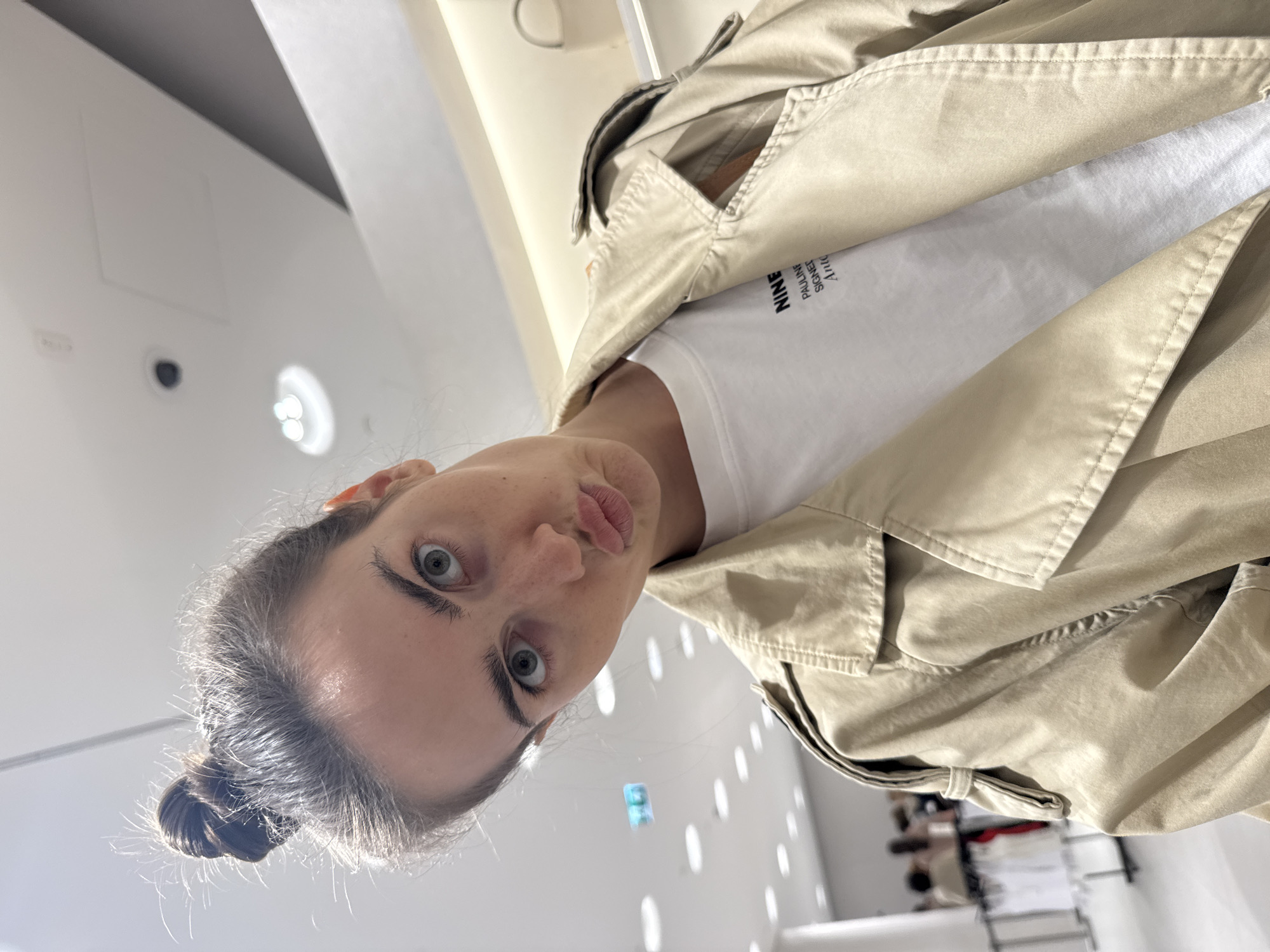 A woman with her hair in a bun, wearing a tan trench coat over a white t-shirt, is photographed from a low angle. The location appears to be inside a brightly lit building, possibly a store, in Unknown, Unknown. Clothing racks are visible in the background.