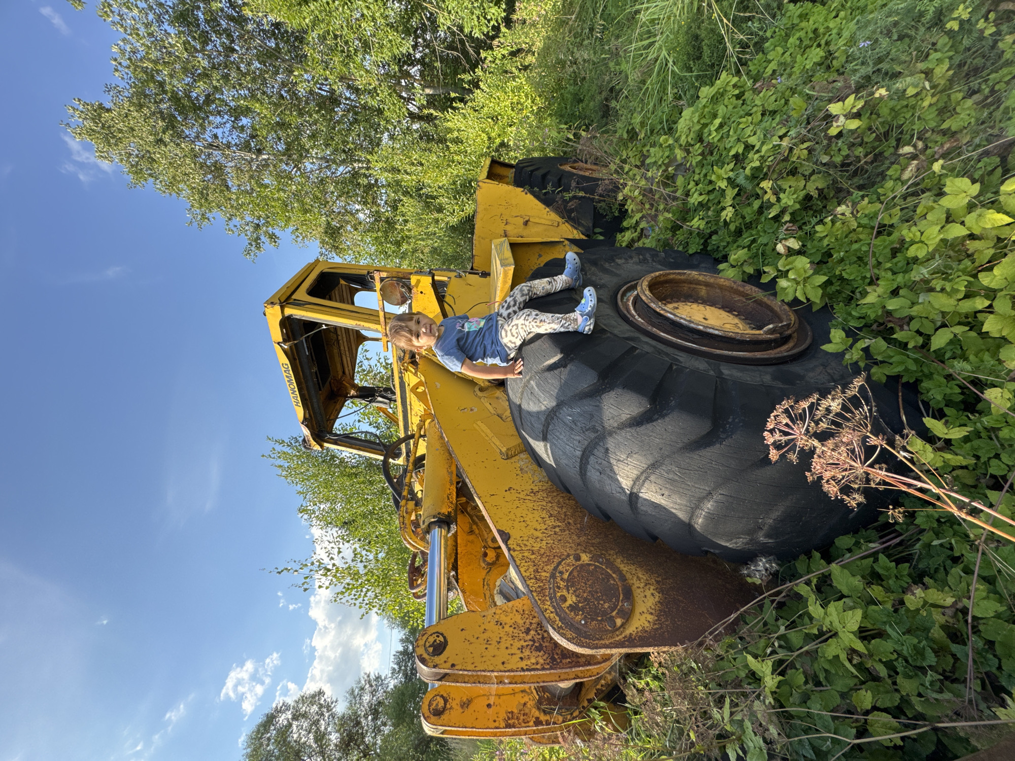 In Innolovo, Russia, a young child sits on the large tire of a yellow, weathered wheel loader. The child is wearing a blue t-shirt with a graphic print and patterned leggings. The wheel loader is parked in a grassy, overgrown area with trees and foliage surrounding it. The sky is blue with scattered white clouds, indicating a sunny day. The machinery appears aged and possibly abandoned, with visible rust and wear. The overall scene captures a moment of childhood exploration in a rustic, natural setting.