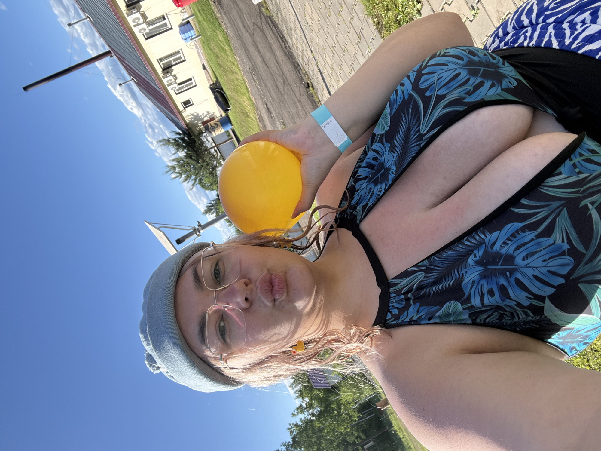 A young woman with blonde hair and glasses is taking a selfie outdoors. She is wearing a light blue beret and a black swimsuit with a blue leaf pattern. She is holding a yellow ball in her right hand and has a blue wristband on her left arm. The background shows a building, trees, and a blue sky with clouds. The location is not identifiable as Birobidzhan, Russia.