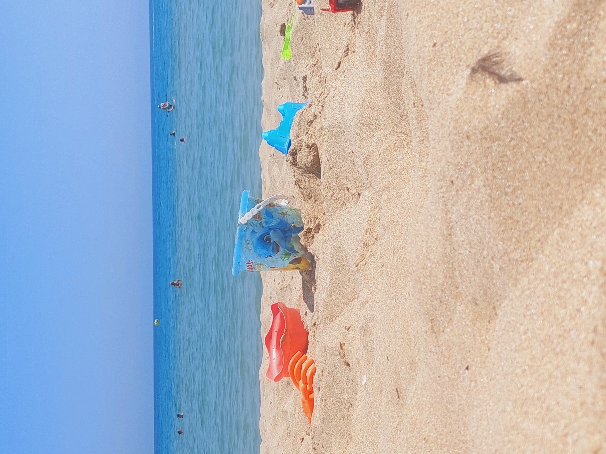 The image depicts a beach scene with the Black Sea in the background. In the foreground, on the sand, are children's beach toys including a red shovel and bucket, and a blue bucket with a dolphin design. A blue sandcastle mold is also visible. Several people are in the water, some swimming and one person appears to be paddleboarding. The sky is clear and blue. This scene is typical of the beaches found in Sozopol, Bulgaria, a popular coastal town known for its sandy beaches and clear waters.