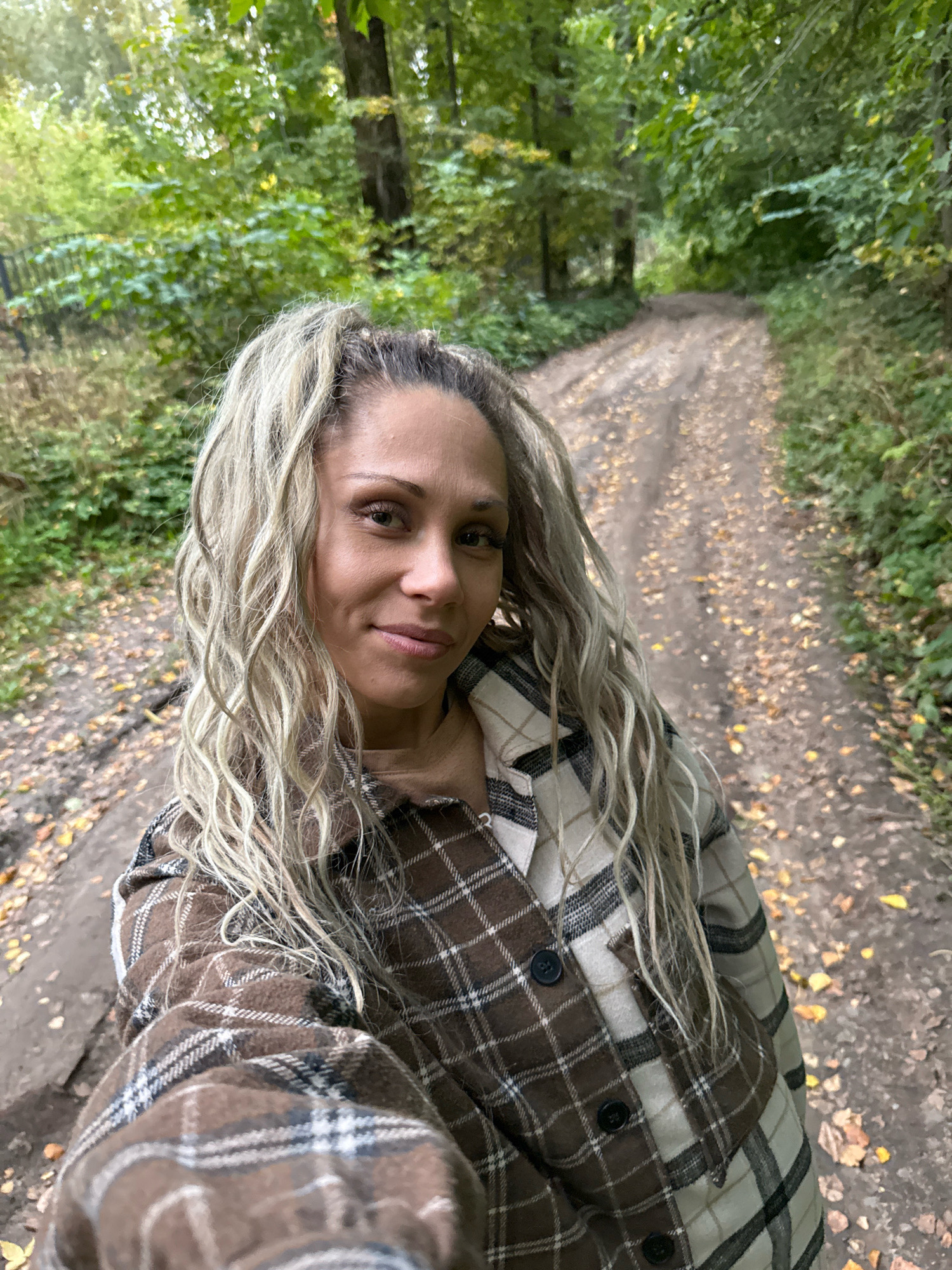 A woman with blonde, wavy hair and prominent facial features takes a selfie on a dirt path in a forest. She is wearing a brown and white plaid shirt. The path is covered with fallen leaves, and trees line both sides of the path. The scene is consistent with an outdoor location in Rusanovo, Russia, during autumn.