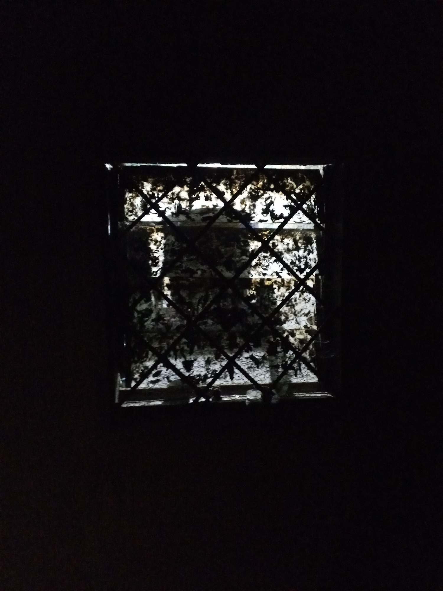A small, square window is centered in the frame against a dark background. The window has a lattice pattern of black bars and is dirty. Outside the window, a light source reflects off of something mottled and unclear. The location is likely Jos, Nigeria, based on the prompt.