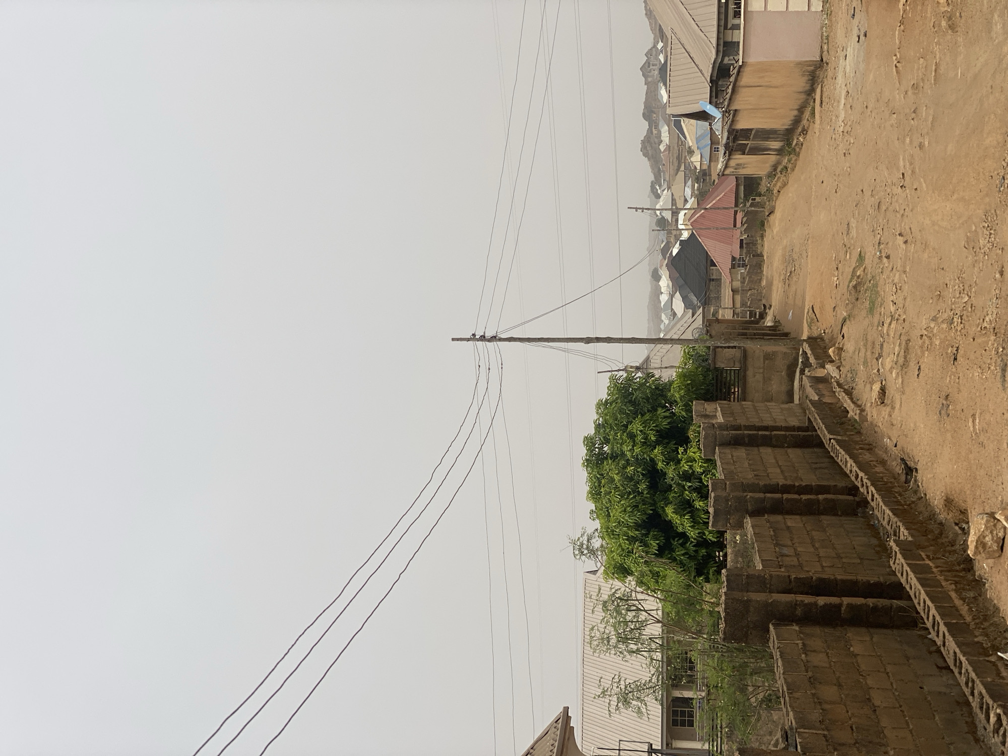 This image depicts a street scene in Barakin Akawo, Nigeria. The view is a slight upward angle, along a dirt road flanked by a low wall constructed of stacked cinder blocks. The road surface is uneven and appears dry. Beyond the wall and road are buildings, varying in height and construction, most with corrugated metal roofs. Utility poles with overhead power lines cut across the sky. The sky is overcast, suggesting a hazy or smoggy atmosphere. No people or animals are visible in the frame. The vegetation appears limited to a few small trees growing among the buildings.