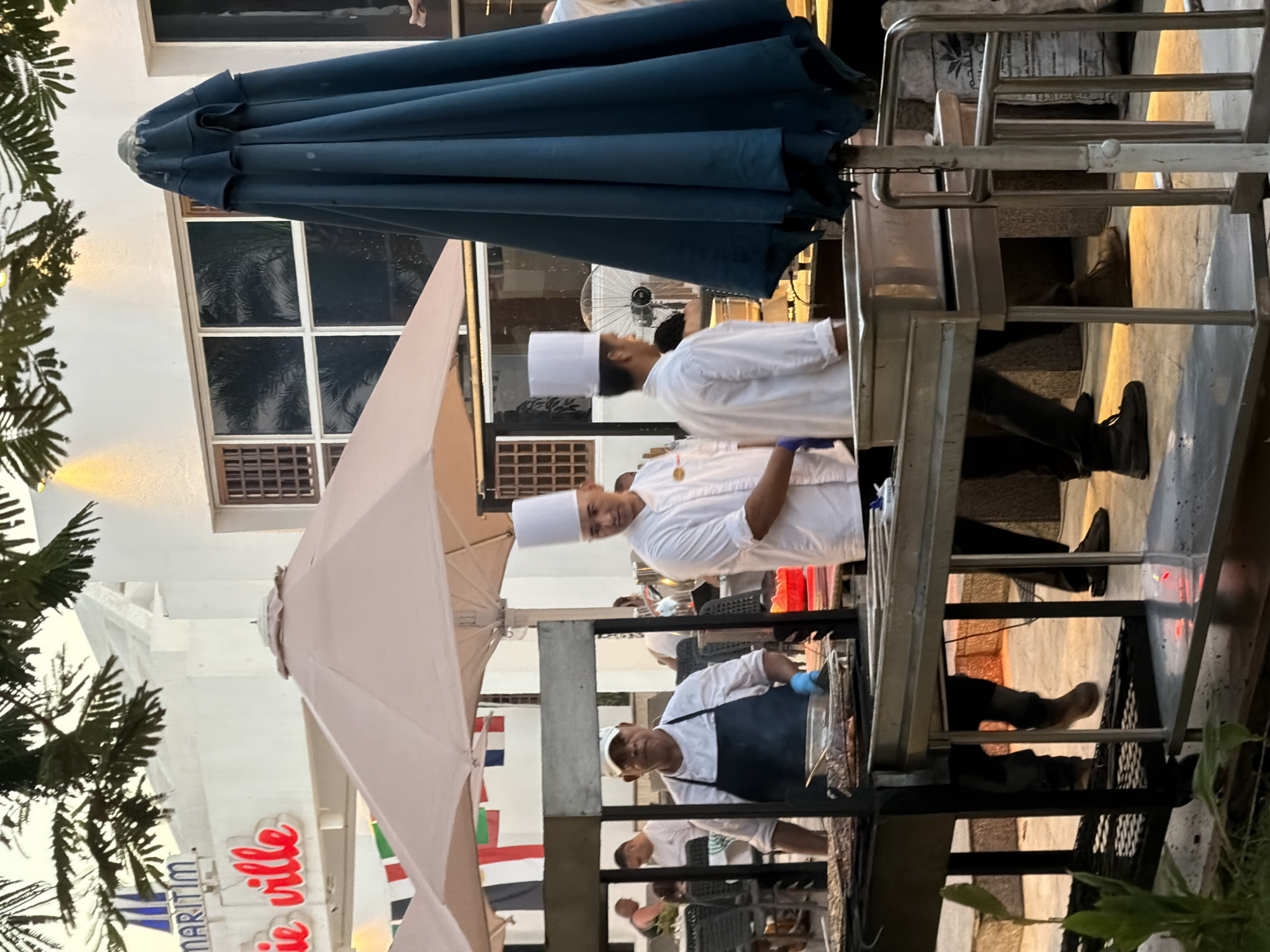 The image captures a lively outdoor dining scene, likely in Sharm El Sheikh, Egypt, given the visible flags and architectural style. The foreground features a professional grilling station, where two chefs in white chef hats and uniforms are actively working. One chef, wearing a dark blue apron and blue gloves, is tending to food on a large grill. The other chef, on the right, appears to be overseeing the operation. Behind them, large umbrellas provide shade for outdoor seating areas, and patrons can be seen dining in the background. The setting is illuminated by soft, warm light, suggesting it might be late afternoon or early evening. The overall atmosphere is one of a well-established restaurant or resort, catering to an international clientele, as indicated by the display of various national flags.