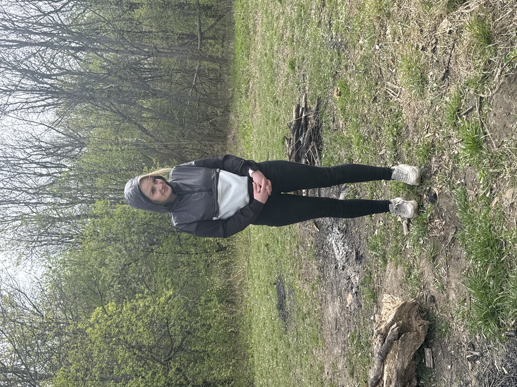 A young woman stands in a grassy area near a burned-out campfire. She is wearing a dark-colored hooded jacket, black pants, and white sneakers. The environment appears to be a wooded area, likely a park or natural area. The ground is covered in grass, dead leaves, and some bare earth. In the background are trees with budding leaves, suggesting springtime. There are remnants of a fire pit with charred wood and ash. It's impossible to determine if the location is Sestroretsk, Russia, without further information or landmarks.
