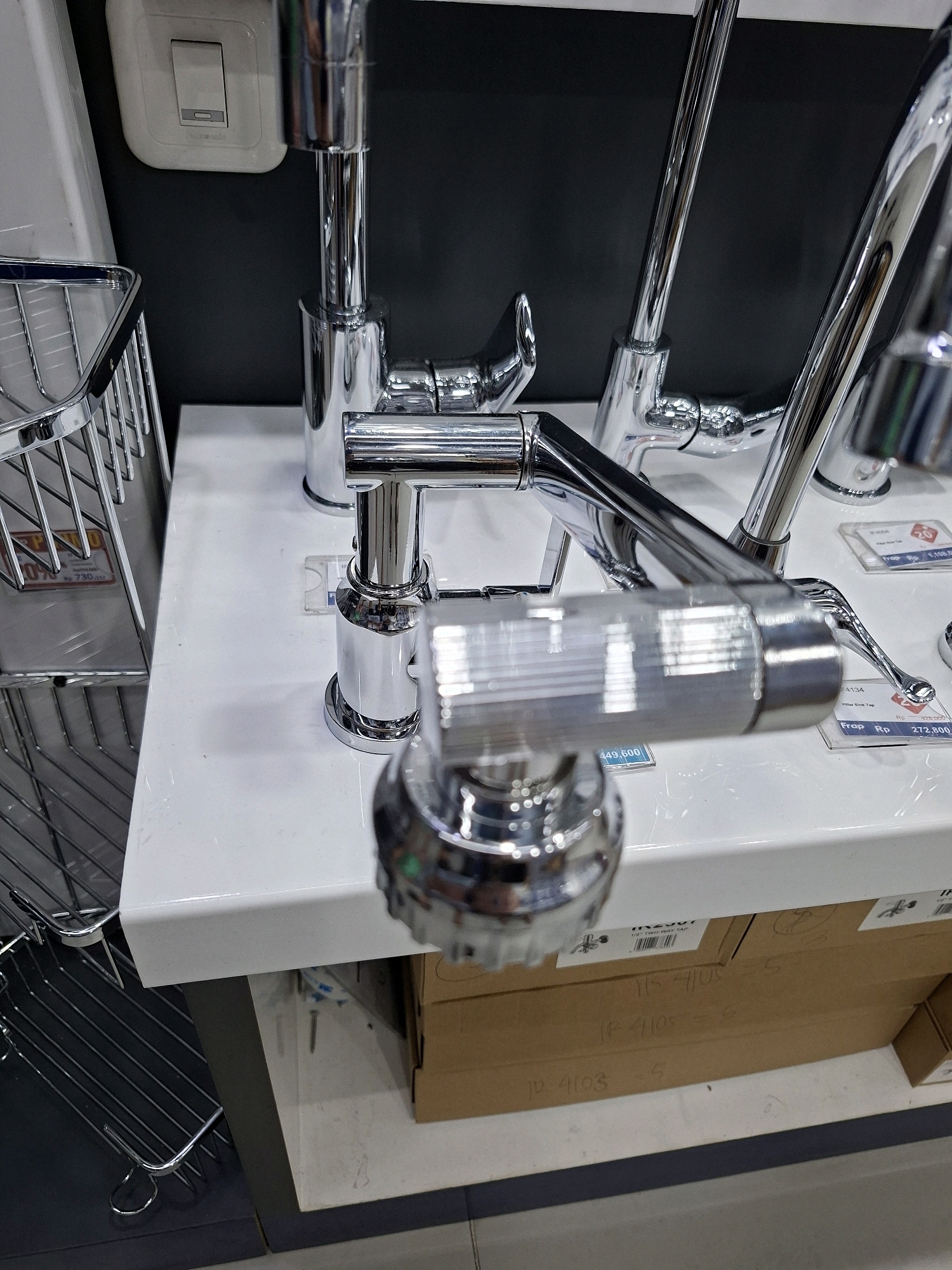 A close-up, slightly overhead shot showcases several chrome faucets and a wire basket on a white countertop. To the left, a chrome wire basket with a red price tag is visible, attached to a dark gray vertical surface. In the background, a white light switch is mounted on a dark gray wall. Several cardboard boxes are stacked beneath the countertop, with handwritten labels. One box has "18 4103" and "5" written on it. Another box has "18 4105" and "5" written on it. The scene appears to be an aisle or display area within a retail store, possibly a hardware or home improvement store, in South Tangerang, Indonesia.