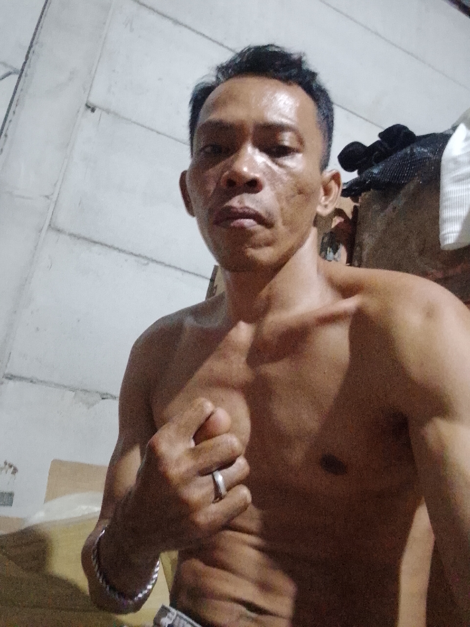 A shirtless man is positioned in a selfie-style photograph. He is wearing a ring on his left hand and a silver bracelet. The background appears to be an indoor setting, with a partially visible wall and other items present. The specific location is in Bukit Ketok, Indonesia.