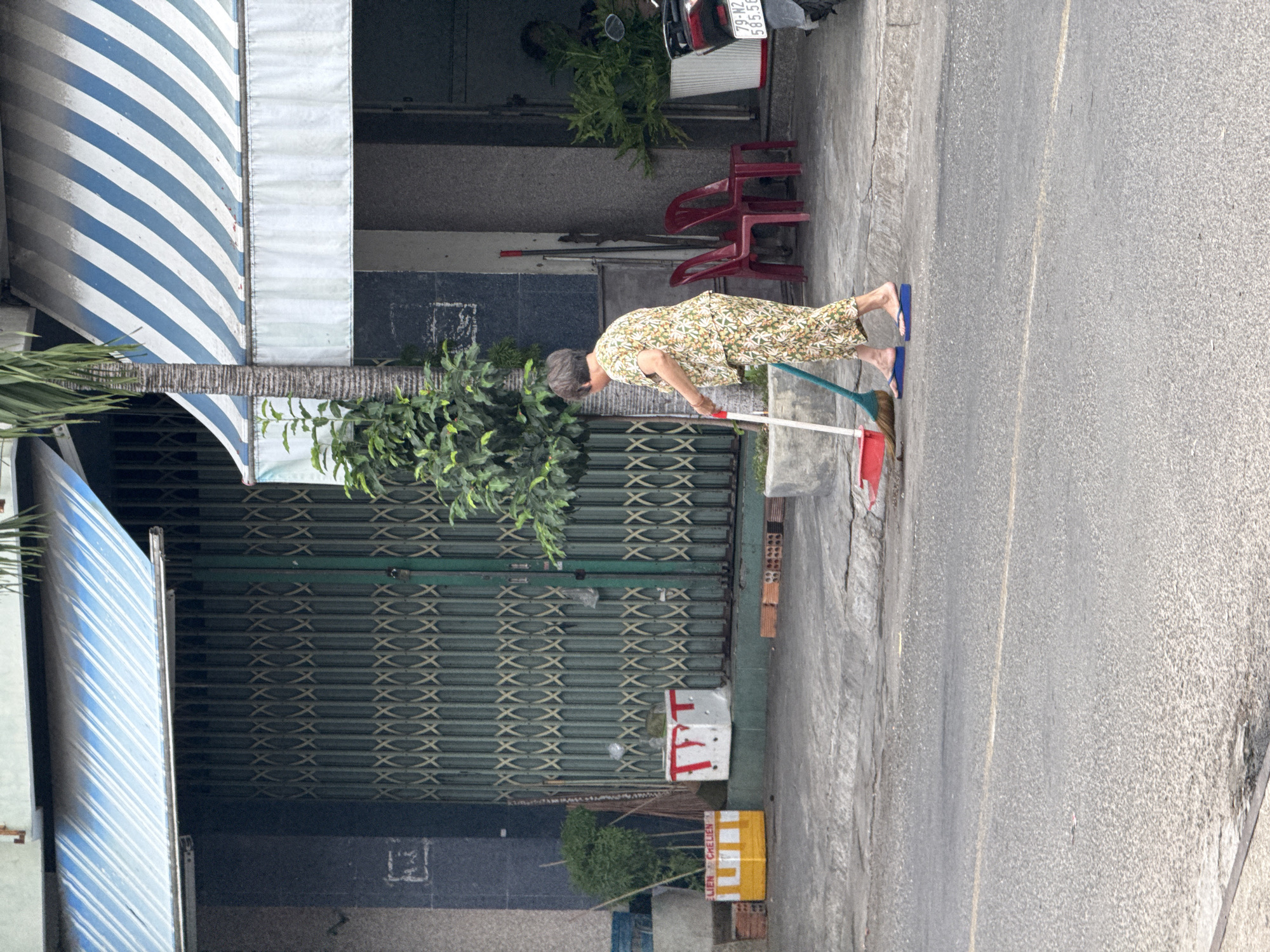 An elderly Vietnamese woman sweeps the sidewalk in front of a building in Nha Trang, Vietnam. She is wearing a floral print pajama set and blue flip-flops. The building has a green metal roll-up gate and a blue and white striped awning. There are two red plastic chairs outside the building, as well as a motorcycle. The sidewalk is made of concrete, and there is a street in the foreground. The scene appears to be a typical street view in a Vietnamese city.
