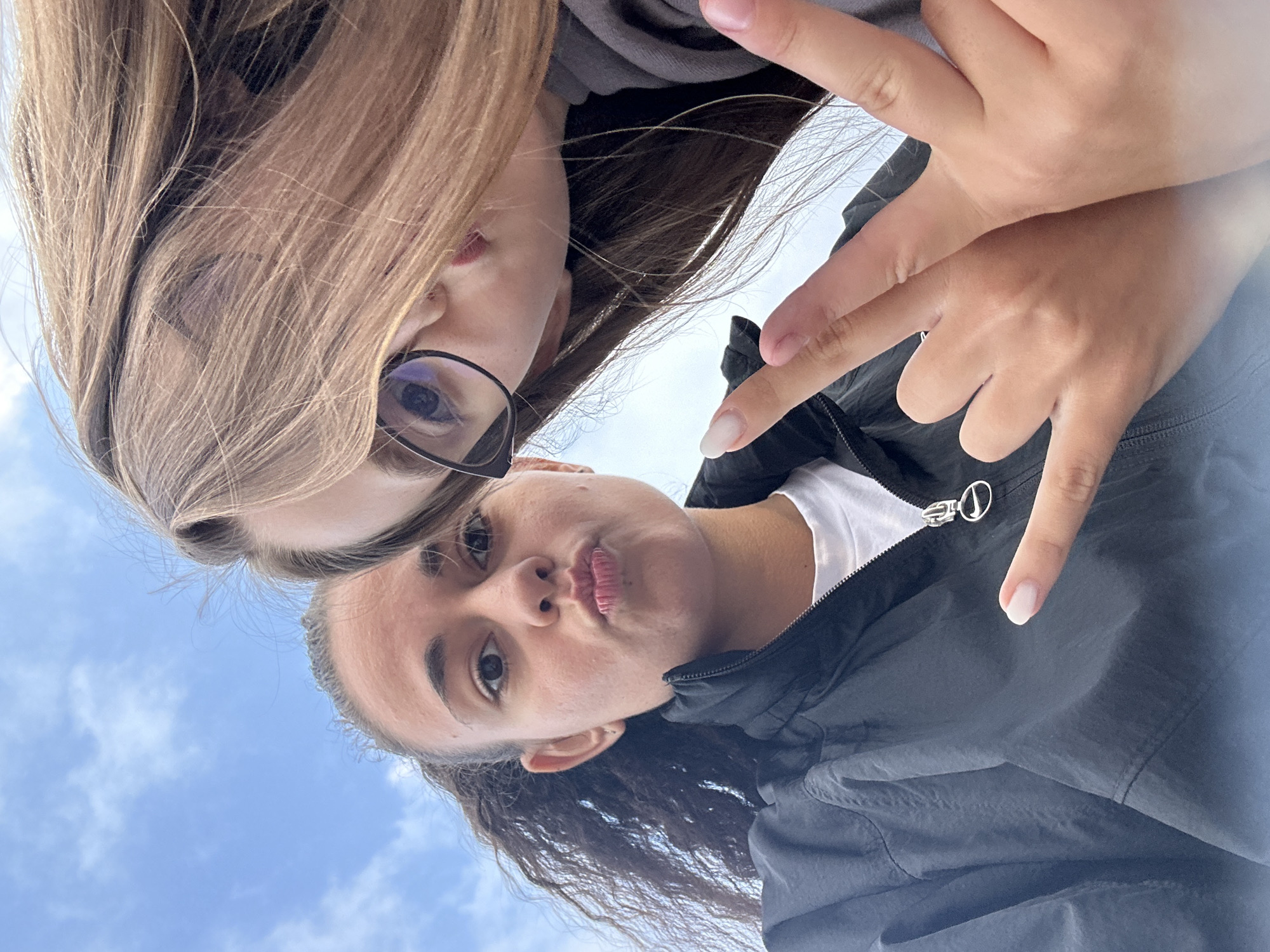 Two young women are captured in a close-up, low-angle selfie against a backdrop of a cloudy sky. The woman on the left has dark, curly hair pulled back in a ponytail and is puckering her lips, wearing a black zip-up jacket over a white t-shirt. The woman on the right, partially obscured by her light brown hair, wears glasses and is leaning towards the camera, her mouth slightly open. Her right hand is raised, displaying two fingers in a peace sign, and her left hand is positioned near her face, also forming a peace sign. The overall atmosphere suggests a casual, outdoor setting. The location is the city of Samokov, Bulgaria, under a partly cloudy sky.