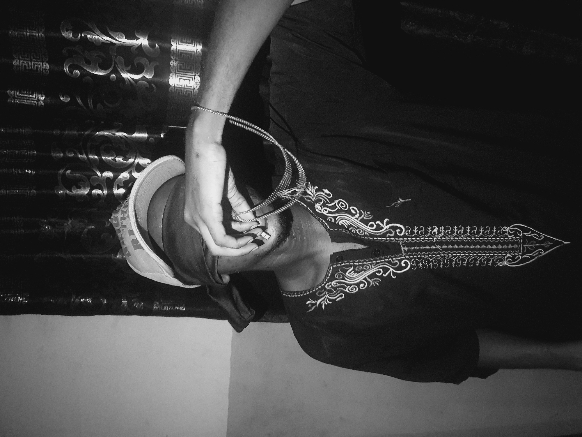 A black and white, low-angle shot shows a person, possibly male, lying on their side. The individual is clad in a dark, embroidered garment. Their head is partially covered with a patterned hat. Their hand, adorned with a bracelet, holds a multi-strand necklace or chain near their face. The background includes a patterned curtain. The location is identified as Lokoja, Nigeria.