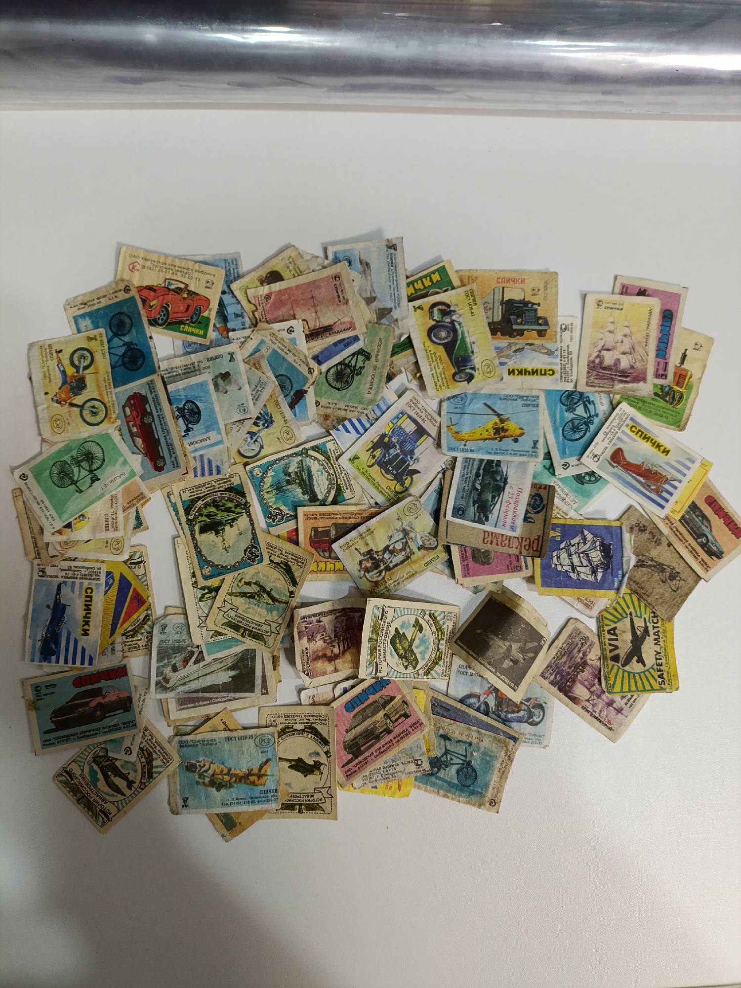 The image shows a collection of vintage matchbox labels scattered on a white surface. The labels are rectangular and feature various illustrations of vehicles, including bicycles, cars, trucks, helicopters, airplanes, and ships. Text in Russian, like "Спички" (matches), is present on many labels. Some labels reference specific Russian cities or factories like "Slobodskoy" and "Belka." One label is marked "AVIA SAFETY MATCH." Many labels are slightly torn and faded, suggesting their age. The background is plain, and the lighting is diffused. It includes city of Chaikovsky, Russia.