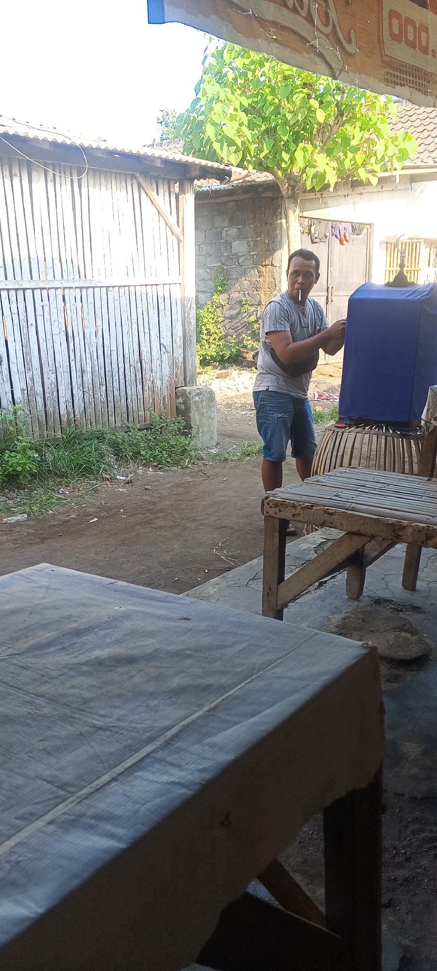 The image depicts a man standing outdoors, smoking a cigarette and appearing to interact with a blue-covered object resting on a woven stand. He is wearing a gray t-shirt, denim shorts, and a black fanny pack. The environment suggests a somewhat rural or semi-urban setting in Lumajang, Indonesia, with a corrugated metal fence, brick walls, and a large tree with green foliage visible. In the foreground, there are two wooden tables, one covered with a dark gray fabric. The lighting suggests it is daytime.