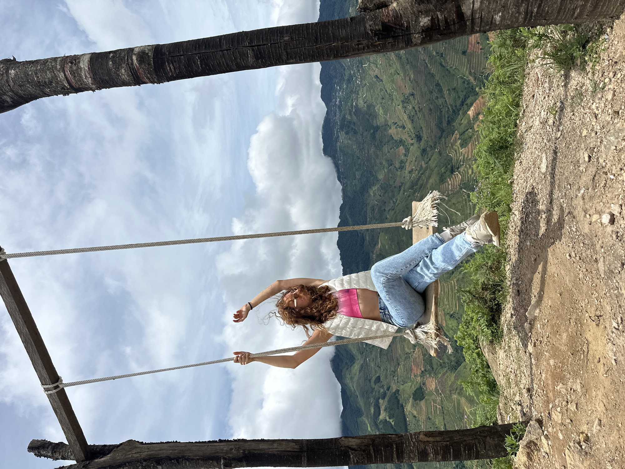 A young woman with curly red hair is sitting on a wooden swing, which is suspended from a rough-hewn wooden beam supported by two dark, textured tree trunks. She is wearing a pink sports bra, a white puffer vest, and light blue jeans. Her arms are raised above her head, and her body is angled as if she is mid-swing. The swing is situated on a grassy, rocky overlook. In the background, a dramatic landscape unfolds with lush green terraced hills and mountains stretching into the distance under a cloudy sky. The scene appears to be in Ta Xua, Vietnam, known for its stunning natural scenery.