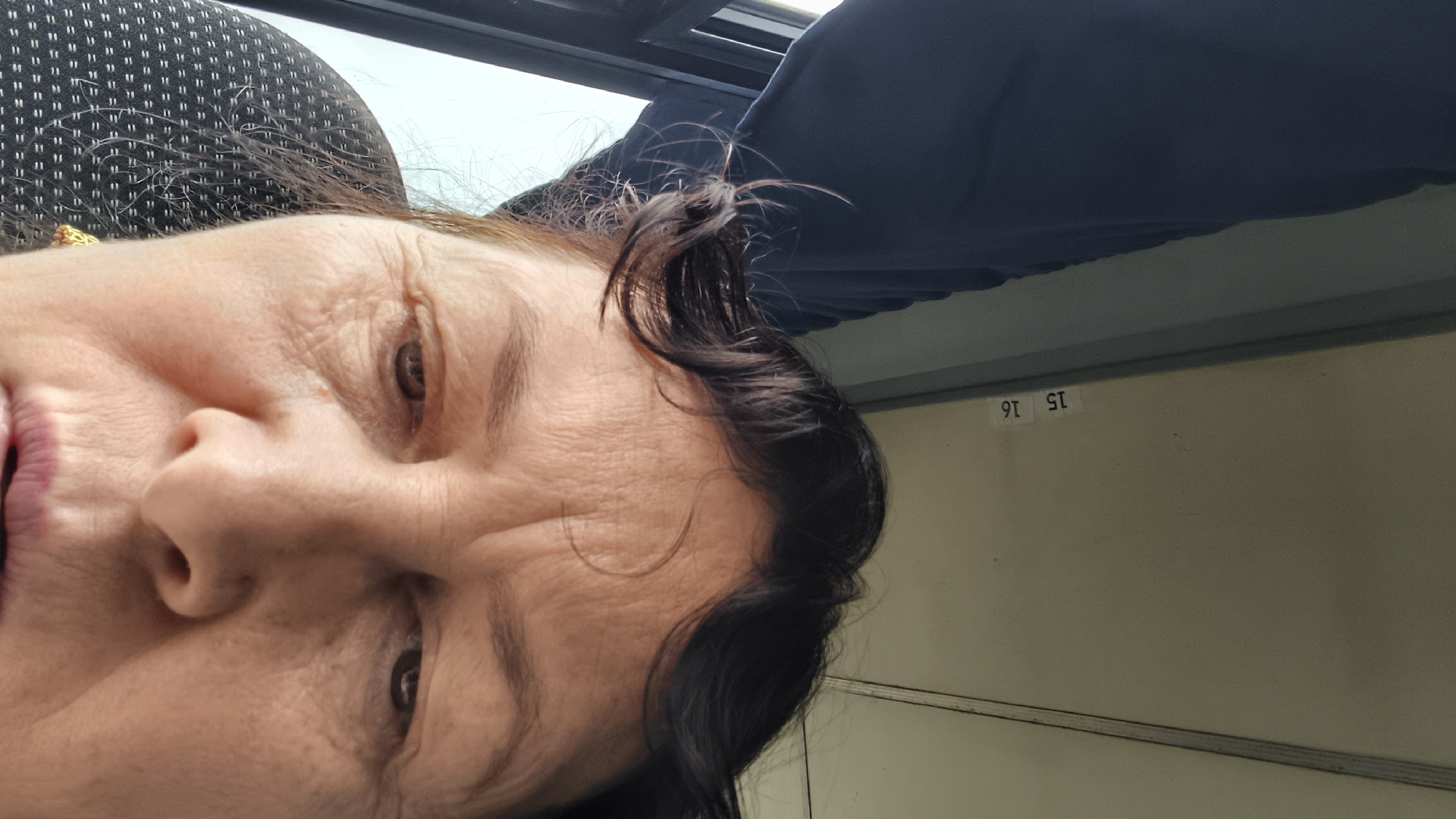 The image is a close-up, low-angle, self-portrait of an older woman. She has dark hair with some strands falling over her forehead and a few wisps are out of place. Her eyes are looking down and to her left and right respectively. Her skin shows visible wrinkles, particularly on her forehead and around her eyes. She is wearing a golden earring in her visible left ear.
The background appears to be the interior of a vehicle, possibly a bus or train, due to the presence of a patterned seat on the left and what looks like a ceiling panel with numbered labels "15" and "16" on the right. A dark blue curtain is visible on the upper left of the image, partially obscuring the window. The lighting suggests it is daytime.
The provided information states the location is Kamyennoye, Russia. However, there are no visual elements in the image that specifically indicate this location. The scene is typical of an indoor transportation setting.