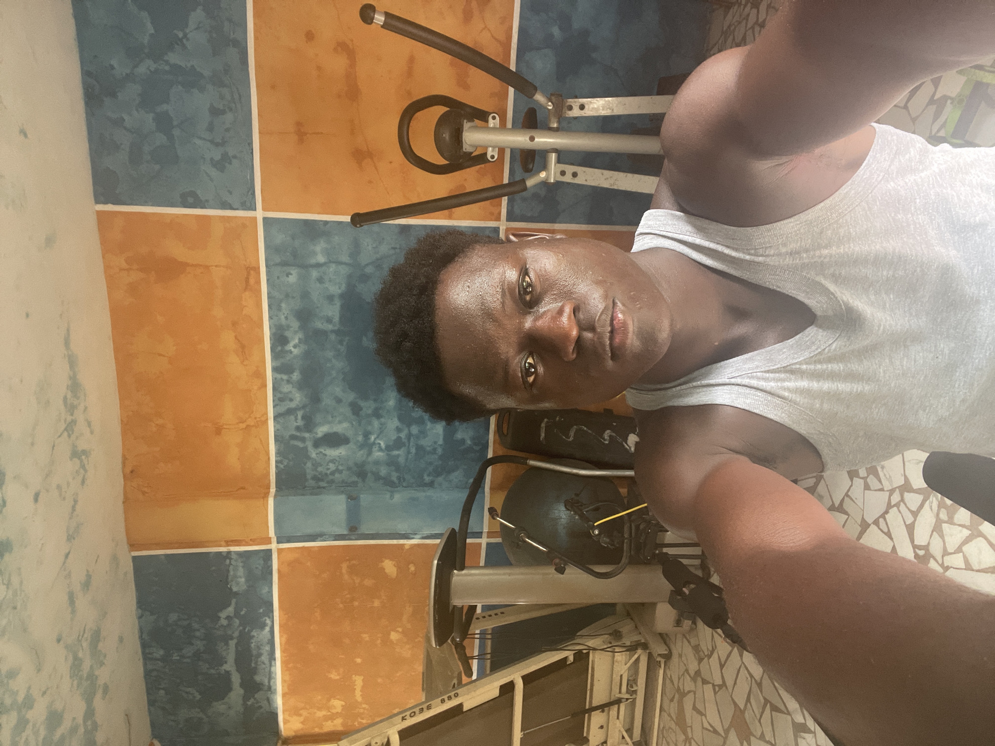 A selfie captures a young man, appearing to be of African descent, indoors in Badagry, Nigeria. The man, with dark skin and short, dark curly hair, is looking directly at the camera with a neutral expression. Sweat is visible on his forehead and face, suggesting recent physical exertion. He is wearing a light grey sleeveless tank top.
The setting appears to be a home gym or an exercise area. Behind the man, a silver and black exercise machine, resembling an elliptical trainer or stepper, is prominent. Another piece of exercise equipment, partially obscured, is visible behind him to the lower right, and a blue exercise ball is also present. The walls are tiled with a striking pattern of alternating orange and blue square tiles, separated by white grout lines. On the far left, another wall is painted in a lighter, mottled color. The floor, visible in the lower right, features a mosaic-like pattern of light-colored, geometric tiles.
Visible text on one of the exercise machines reads "KOBE 680". The lighting suggests it is daytime, possibly indoors with natural light or bright artificial lighting. The overall impression is of a person documenting their workout session.
