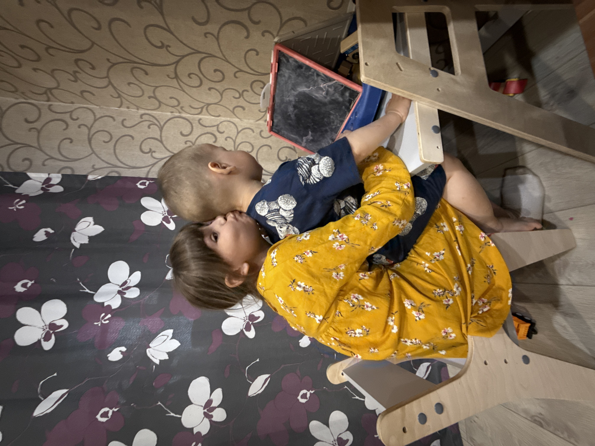 Two children are lying on a floral-patterned surface, possibly a bed or play mat, in what appears to be an indoor setting. The younger child, with light-colored hair, is wearing a dark blue shirt with white patterns. The older child, with brown hair, is wearing a yellow, floral-patterned dress and appears to be hugging the younger child. A wooden climbing structure is partially visible in the right side of the frame, along with a chalkboard in the background. The location is Rusanovo, Russia.