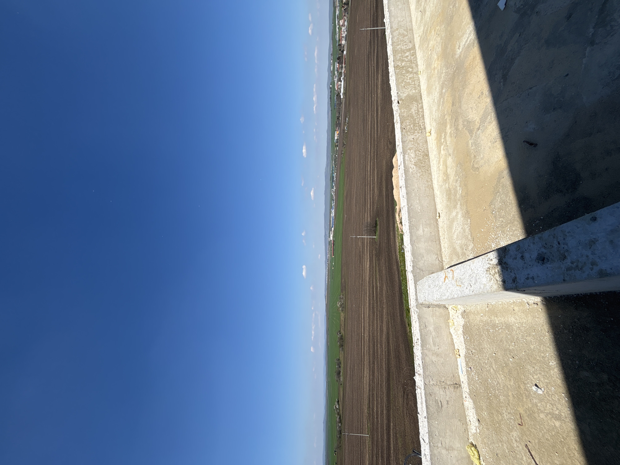 The image appears to be taken from a high vantage point, possibly a building or structure under construction, overlooking a rural or agricultural landscape. The sky occupies a significant portion of the left side of the image, displaying a clear blue color with scattered clouds. A field, primarily of dark brown color, extends across the middle portion of the image, suggesting cultivated land. Further in the distance, a strip of greenery can be seen, bordering a line of buildings that suggest a settlement or town. The right side of the image consists of concrete structures, seemingly part of the building from which the image was taken. The presence of shadows on the concrete indicates a strong source of light, presumably the sun. Given the context of Burgas, Bulgaria, the image could depict the view from a building on the outskirts of the city, overlooking nearby agricultural fields and a small town or village. It is impossible to determine any interactions from the photo.