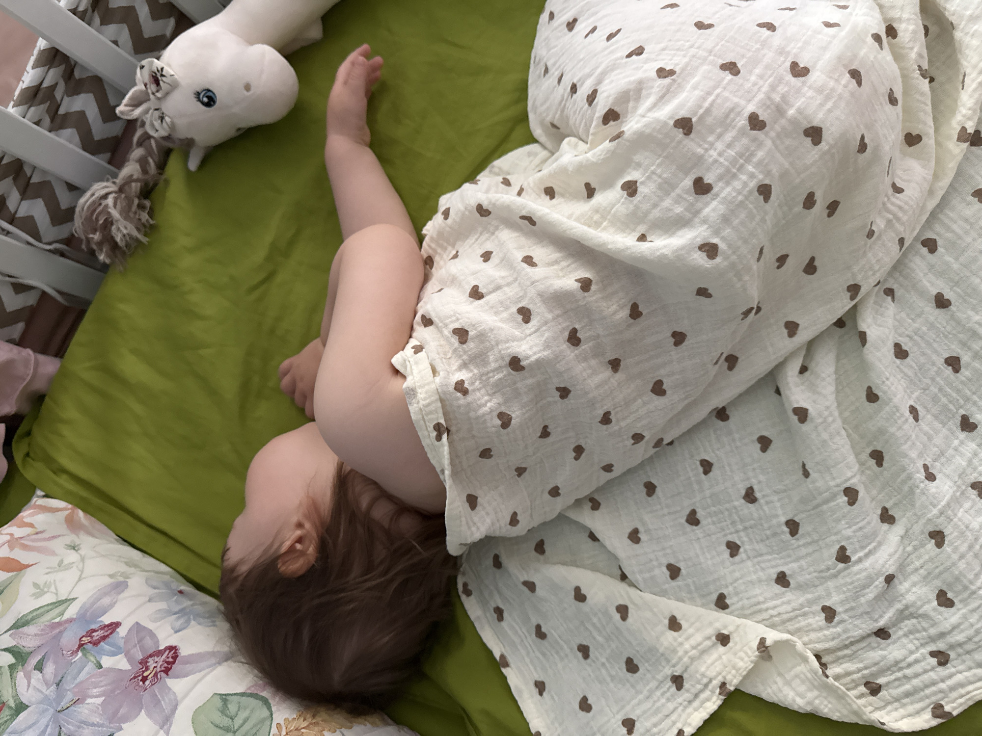 A baby sleeps on a green mattress in a crib. The baby is covered by a white blanket with brown hearts. A white unicorn toy is next to the baby. The crib has white bars and a brown and white chevron pattern. A floral pillow is on the left side of the image. The scene appears to be in a bedroom.