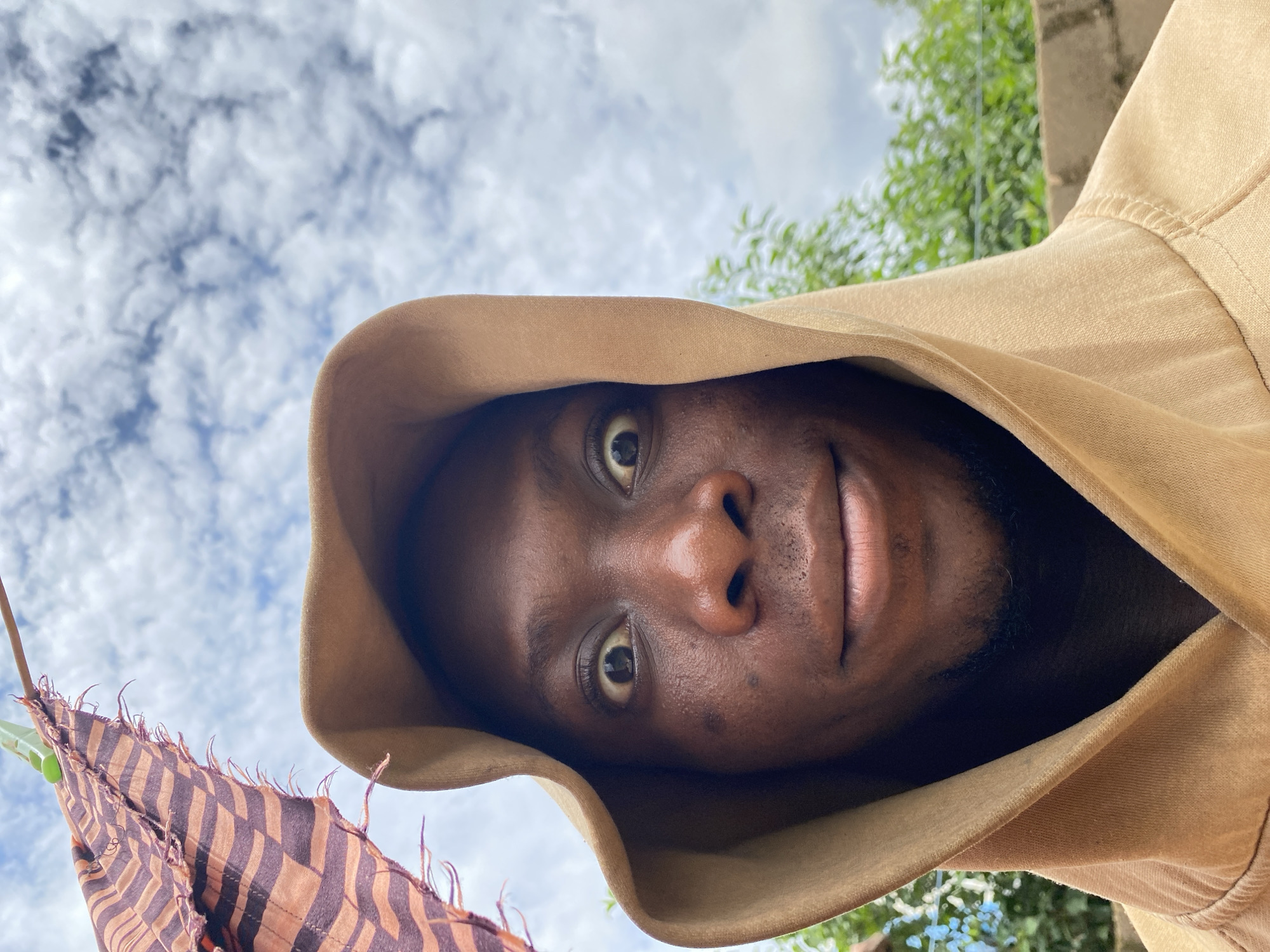 A close-up, low-angle, eye-level outdoor selfie shows a Black male wearing a tan hooded sweatshirt, with the hood pulled up around his face. He has wide eyes and a slight smile. In the upper left corner of the frame, a patterned fabric, possibly a scarf or cloth, hangs from a clothesline. The background consists of a cloudy sky and out-of-focus green foliage, suggesting a natural or semi-urban environment. Based on the provided context, this image was taken in Bukuru, Nigeria.