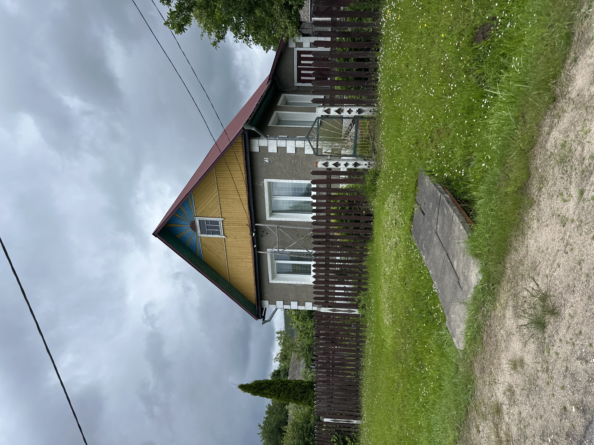 The image depicts a grey, two-story house with a red roof and yellow siding under a cloudy sky. A dark brown wooden fence encloses the property, with a small, ornate metal gate in the center. The yard is covered in green grass with scattered white and yellow wildflowers. A concrete structure, possibly a ramp or platform, is visible in the foreground. A tall, slender evergreen tree stands to the left of the house, and other trees and greenery are visible in the background. The house has several windows, and what appears to be a house number "73" is visible on the exterior wall. The scene suggests a rural or suburban residential setting in Крэўскі сельскі Савет, Belarus. There are no people or animals visible in the image.
