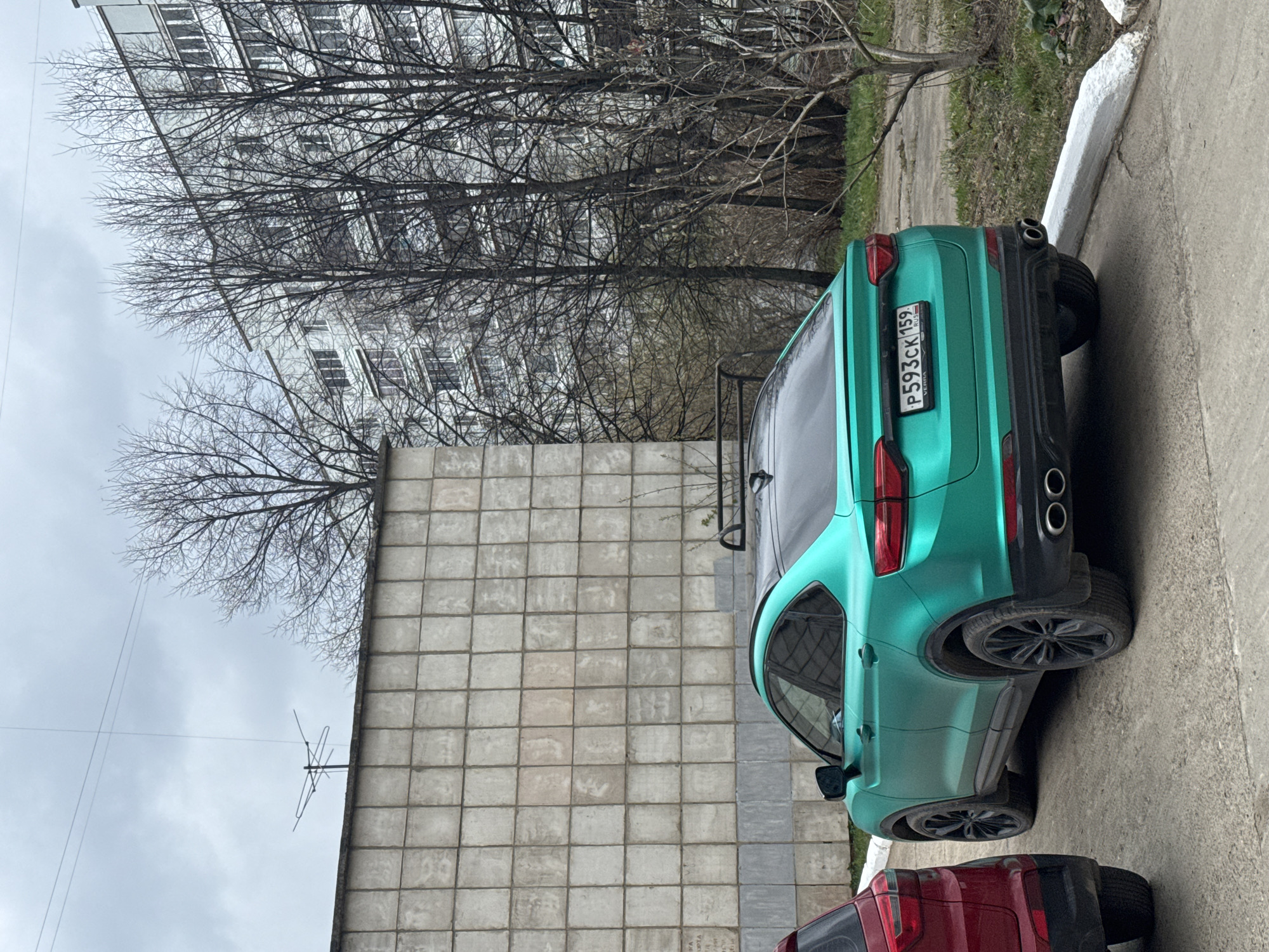 The image, taken in Chaikovsky, Russia, shows a brightly colored teal/green car parked on a paved surface next to a building and bare trees. The car is a Geely model, likely an SUV or crossover, with a black roof and black wheels. Its rear license plate, partially obscured by dirt, reads "P593CK 159 RUSSIA". Below the license plate, the text "GEELY" is visible, along with "MONJARO" and "VERNA". The vehicle features dual exhaust pipes. The rear of a red car is partially visible in the foreground, parked behind the green car.
The setting appears to be an urban or residential area. To the left of the green car, there's a light-colored building with a facade made of large square concrete blocks or tiles. Above this building and in the background, numerous bare deciduous trees with intricate branch patterns are visible, suggesting late autumn, winter, or early spring. Through the branches, another multi-story building with many windows can be seen. The sky is overcast and grey. An old-fashioned television antenna is visible on the roofline of the closer building. The ground consists of grey asphalt or concrete paving, with some patches of dry grass and dirt. A white painted curb runs along the edge of the parking area. No people are visible, and the cars appear to be stationary. The overall atmosphere is muted due to the overcast sky and lack of greenery.