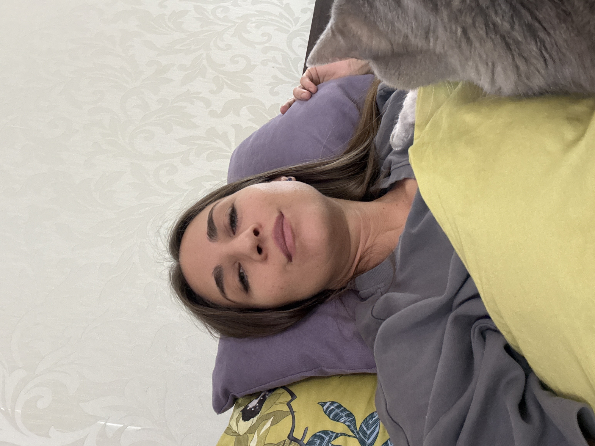 The image presents a woman reclining on a bed. She rests on a purple pillow placed on a bed with a patterned yellow and green cover. She is wearing a grey sweatshirt. A grey cat is near her head. The wall behind the bed has a light patterned wallpaper. The location is in Zanevka, Russia.